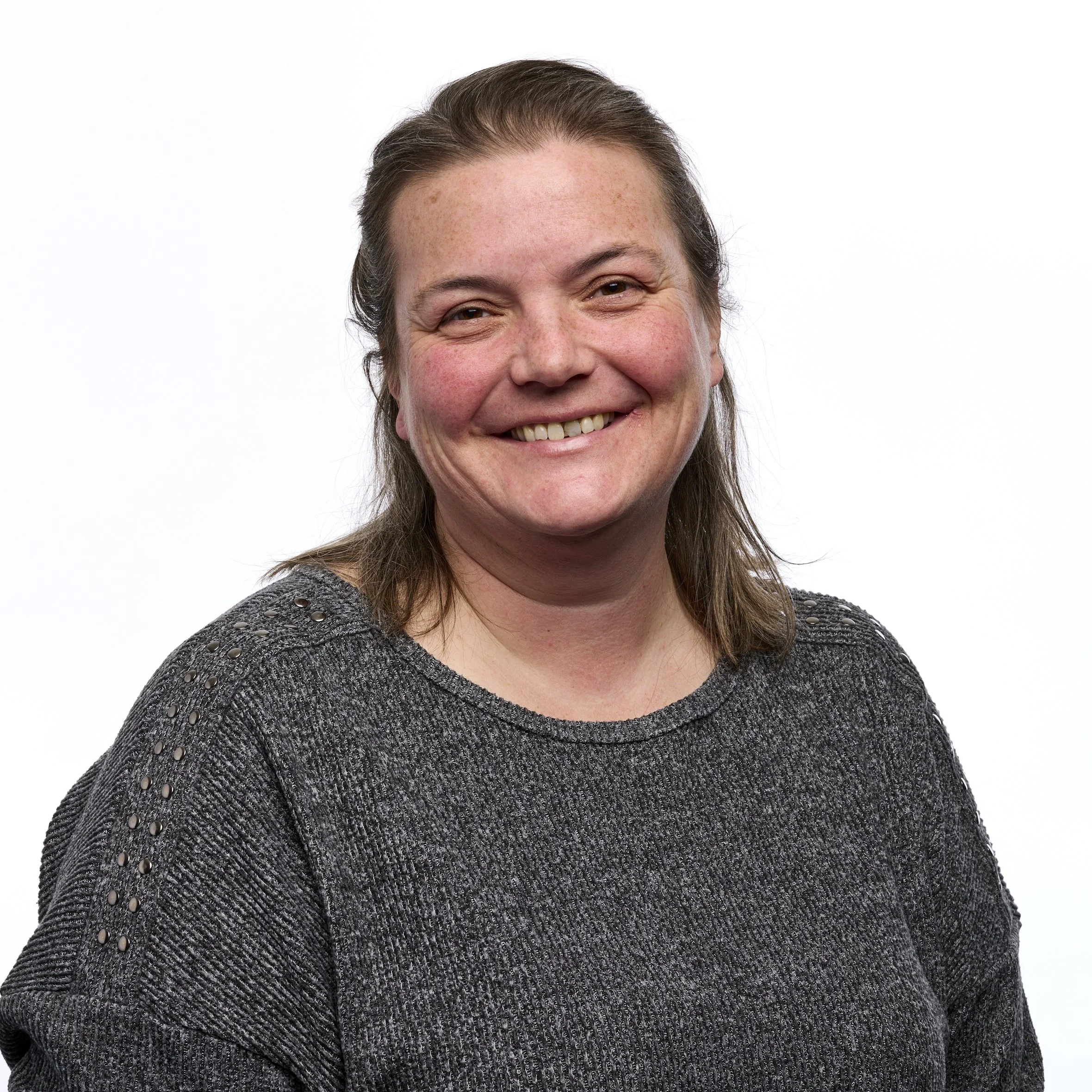 A smiling woman with shoulder-length brown hair wearing a dark gray sweater with studded details on the shoulders, standing against a plain white background.