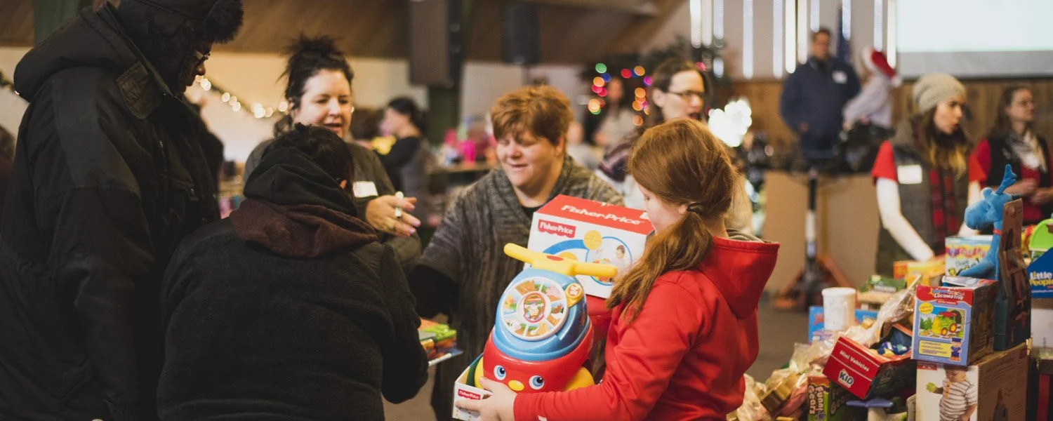 Children and adults at a Christmas charity event, exchanging gifts with a young girl holding a Fisher-Price toy, surrounded by holiday decorations and toy donations.