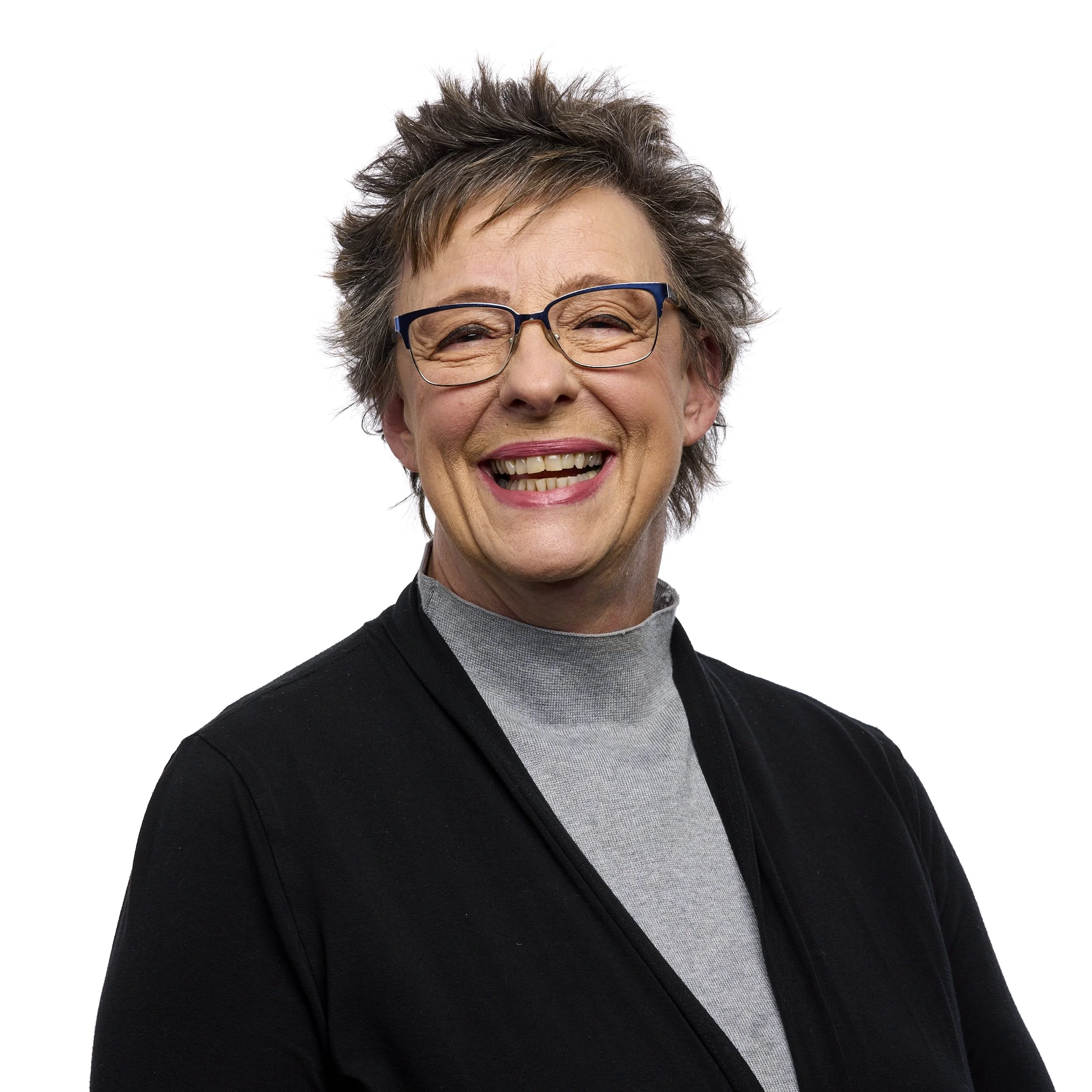 A smiling older woman with short, gray hair, wearing glasses, a black cardigan, and a gray turtleneck shirt.