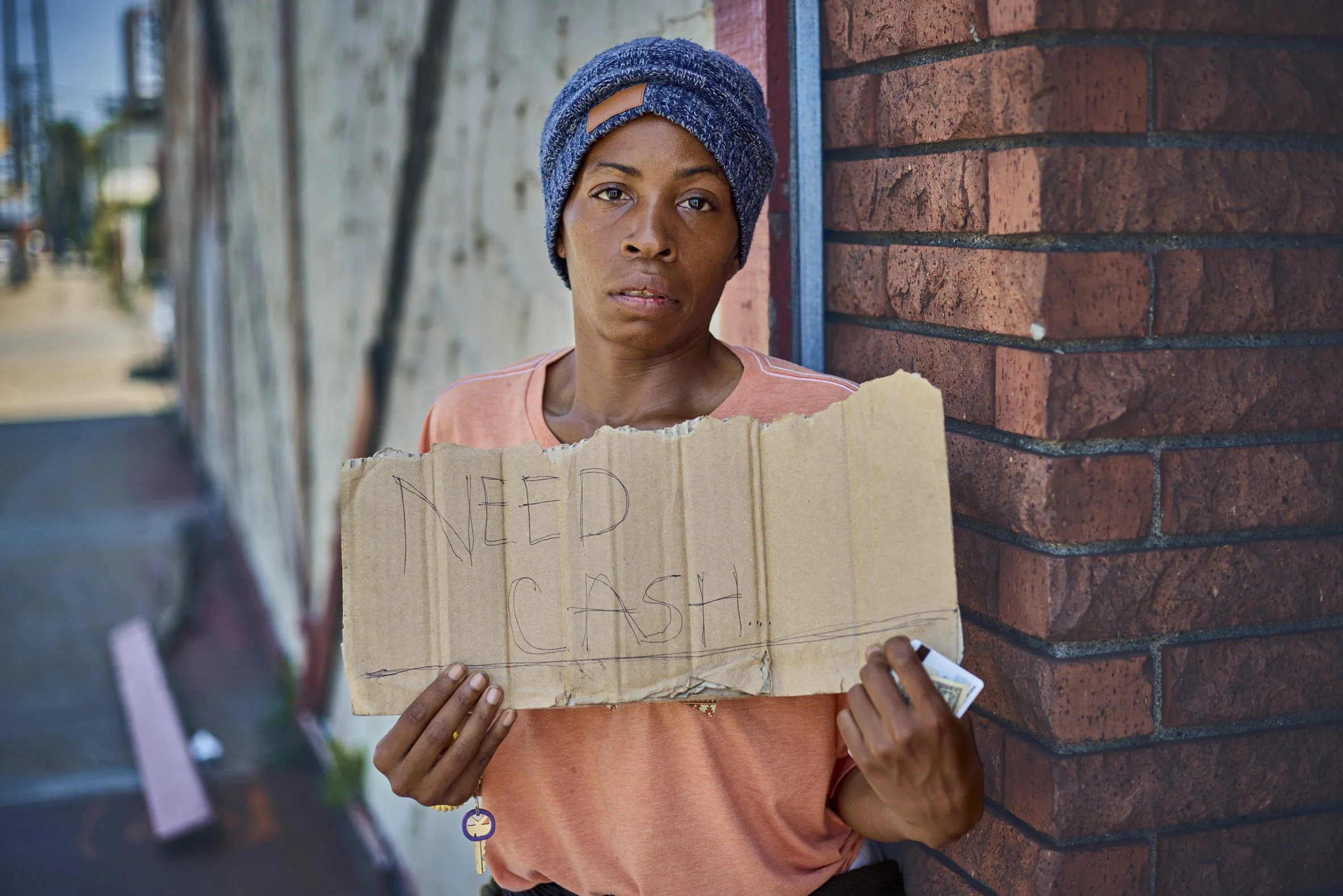 A woman with a blue headwrap and a peach-colored shirt holding a torn cardboard sign that reads 'NEED CASH' in black marker, standing next to a brick wall on a city street.