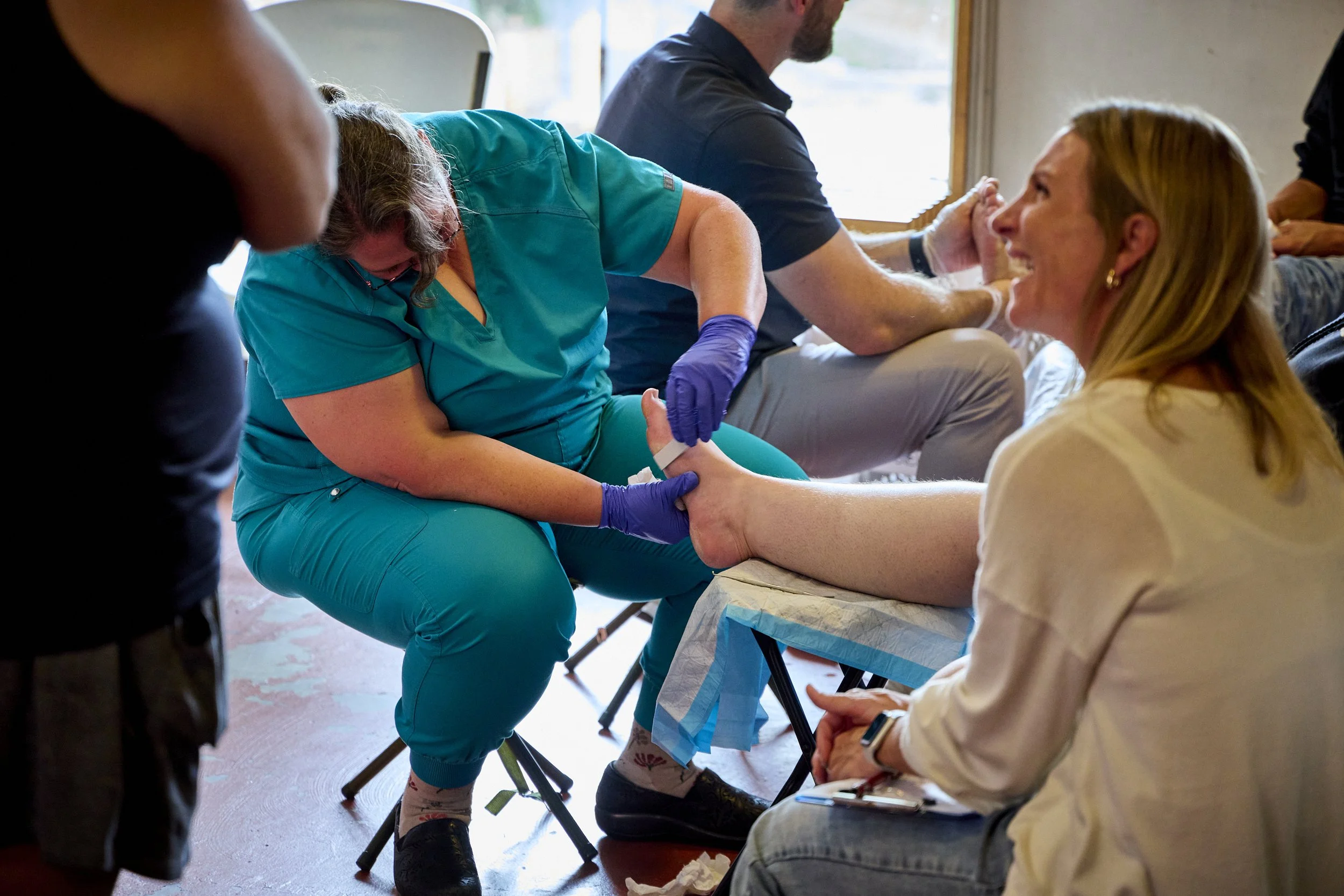 A nurse in teal scrubs with purple gloves helps a woman at a blood drive. The woman is seated with her arm extended, smiling as she donates blood, and a health professional monitors the process. In the background, other donors are seated.
