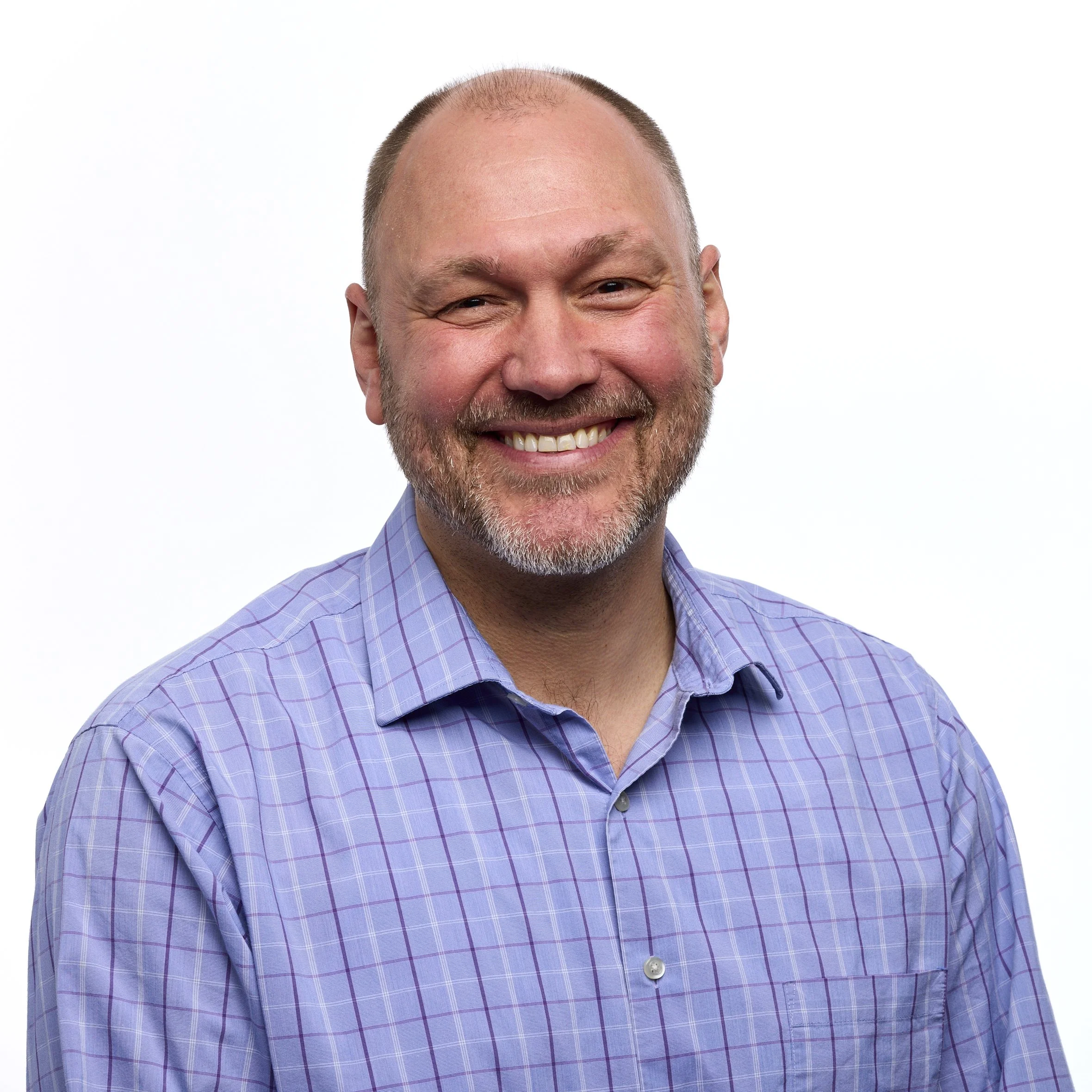 A smiling middle-aged man with a beard and receding hairline wearing a purple checkered shirt against a plain white background.
