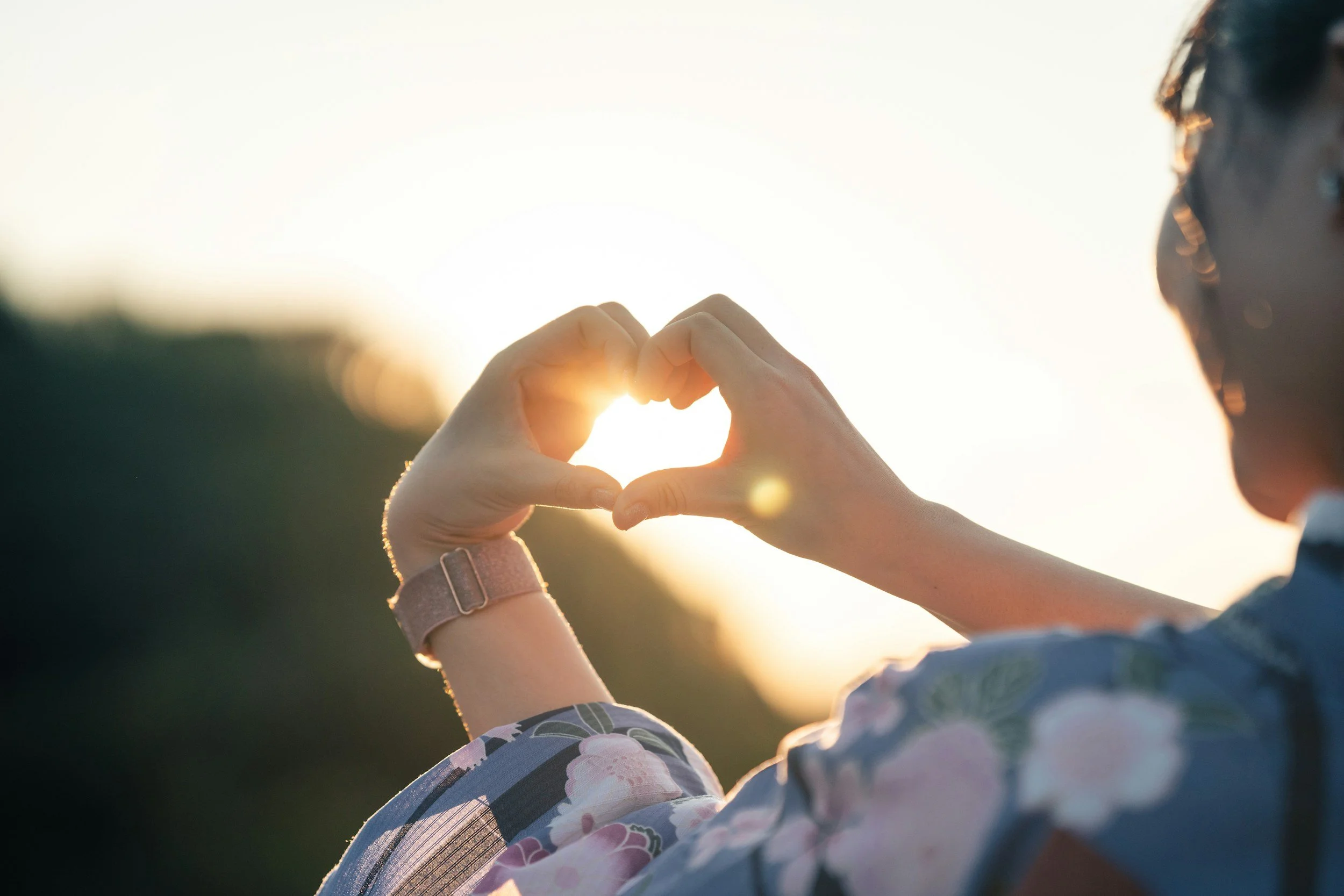 Person forming a heart shape with their hands against a bright sunset background.