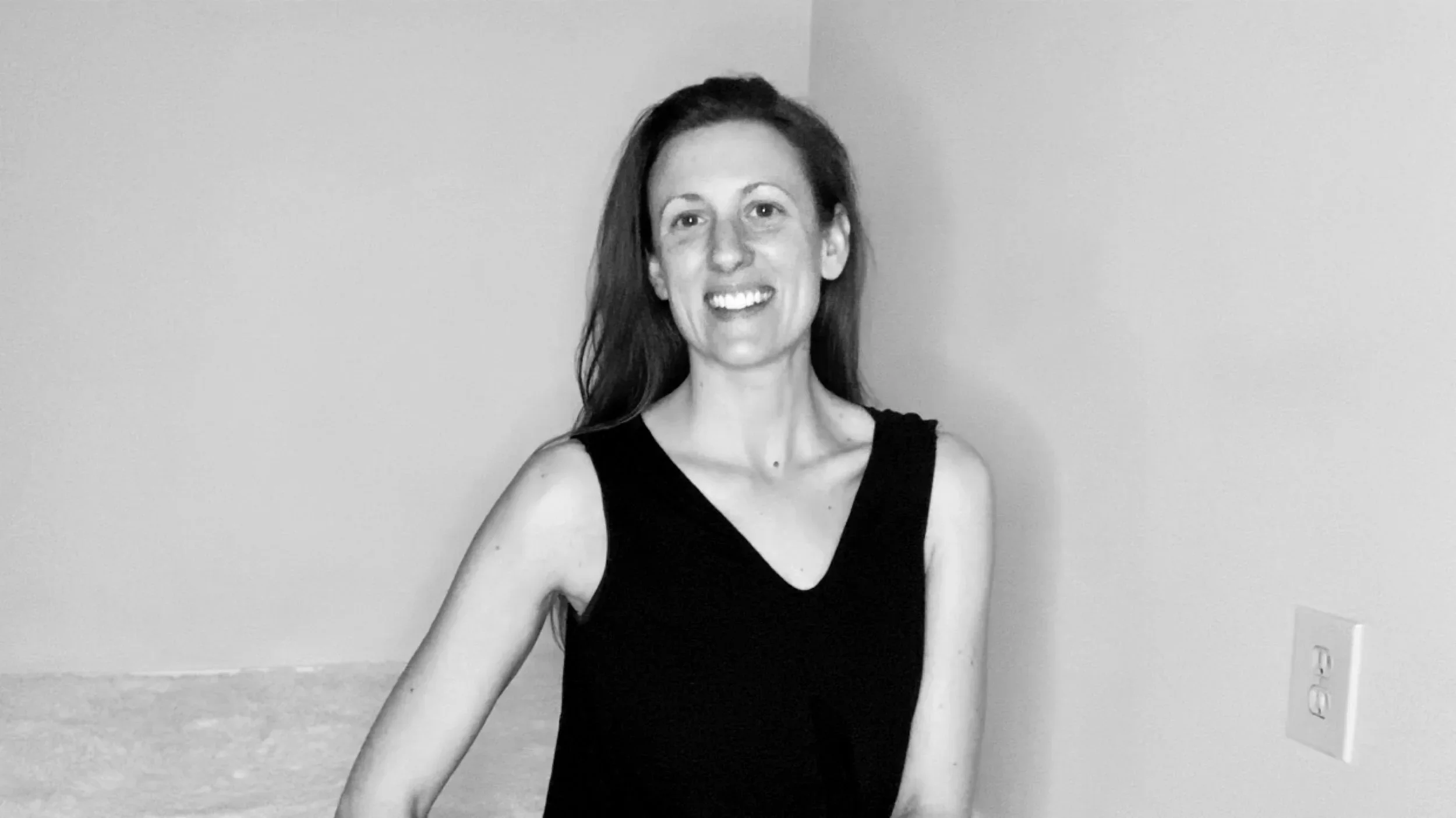 A woman with long hair, smiling, wearing a black sleeveless top, standing indoors against a plain wall.