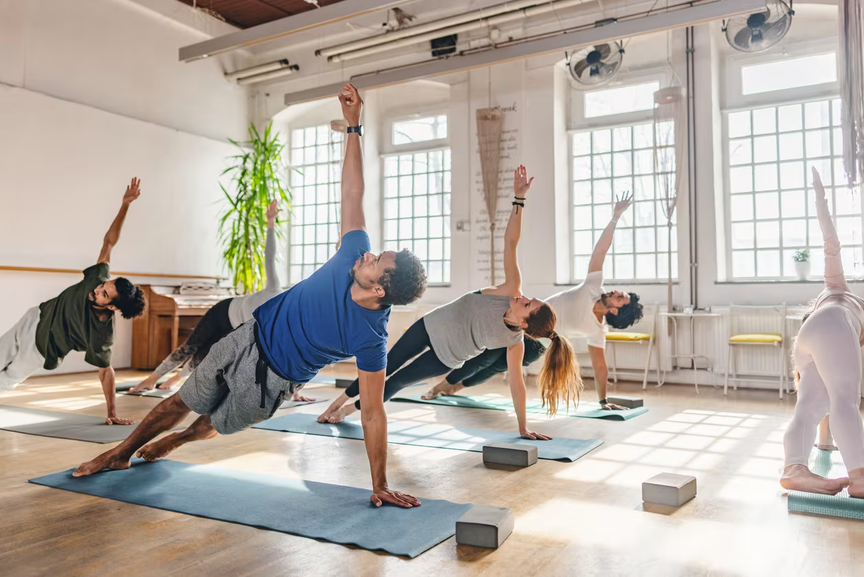 Group of people practicing yoga in a bright, spacious studio, performing side plank pose on yoga mats with yoga blocks nearby.