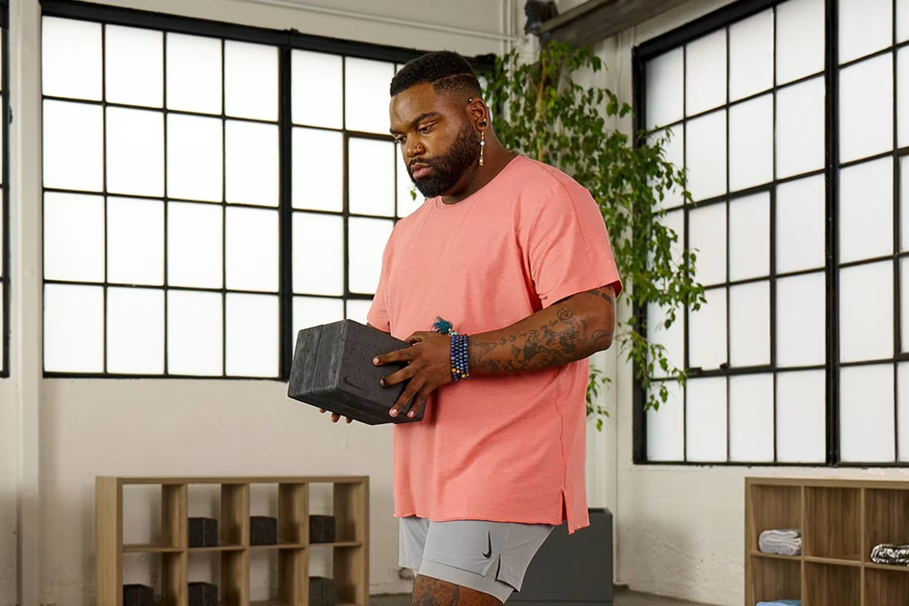 A man with tattoos, wearing a pink t-shirt and gray shorts, holding a black weight in a fitness studio with large windows and natural light.