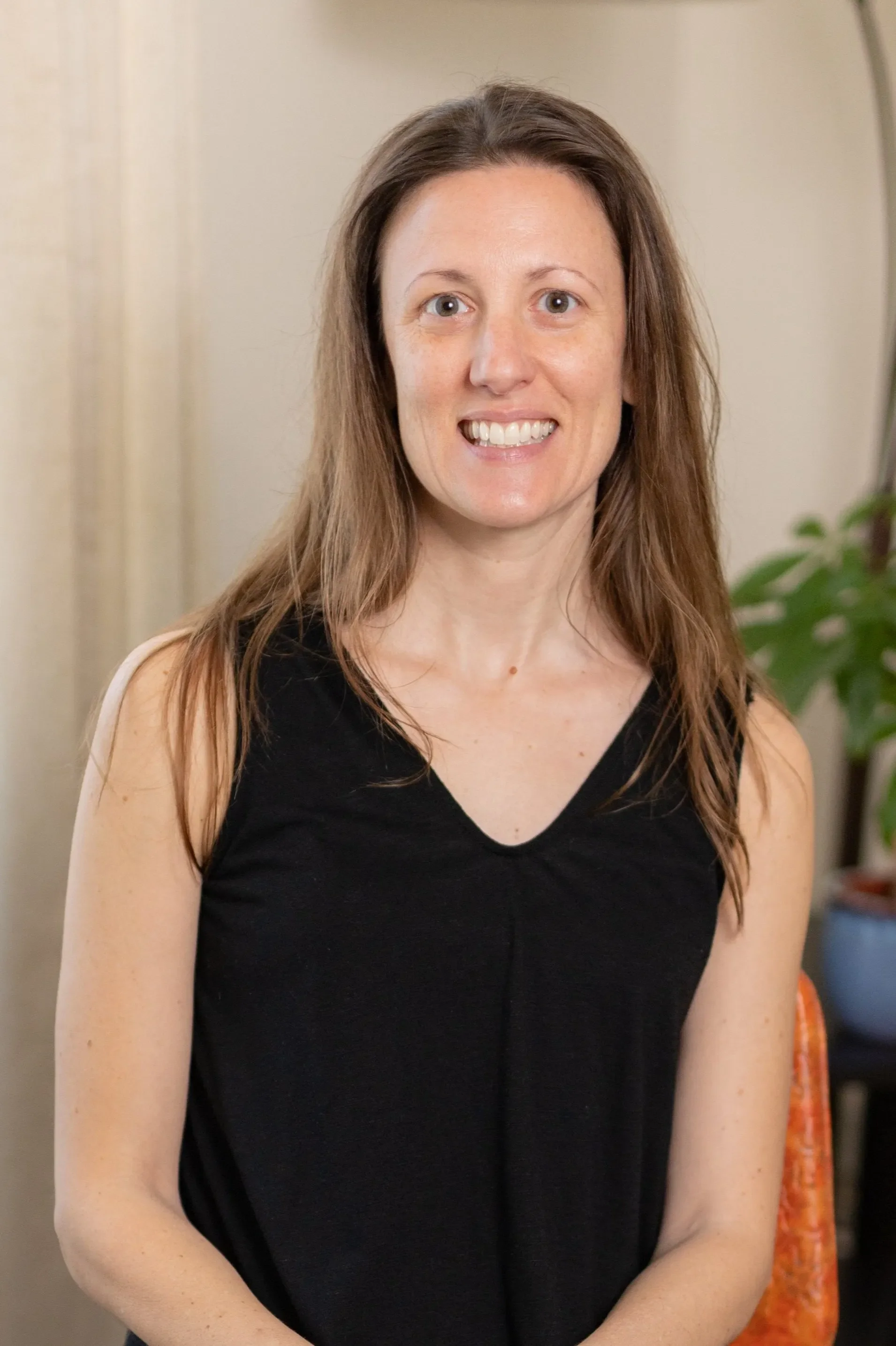 Robyn Gaillard wearing a black sleeveless top, smiling, sitting on an orange chair with a potted plant in the background.