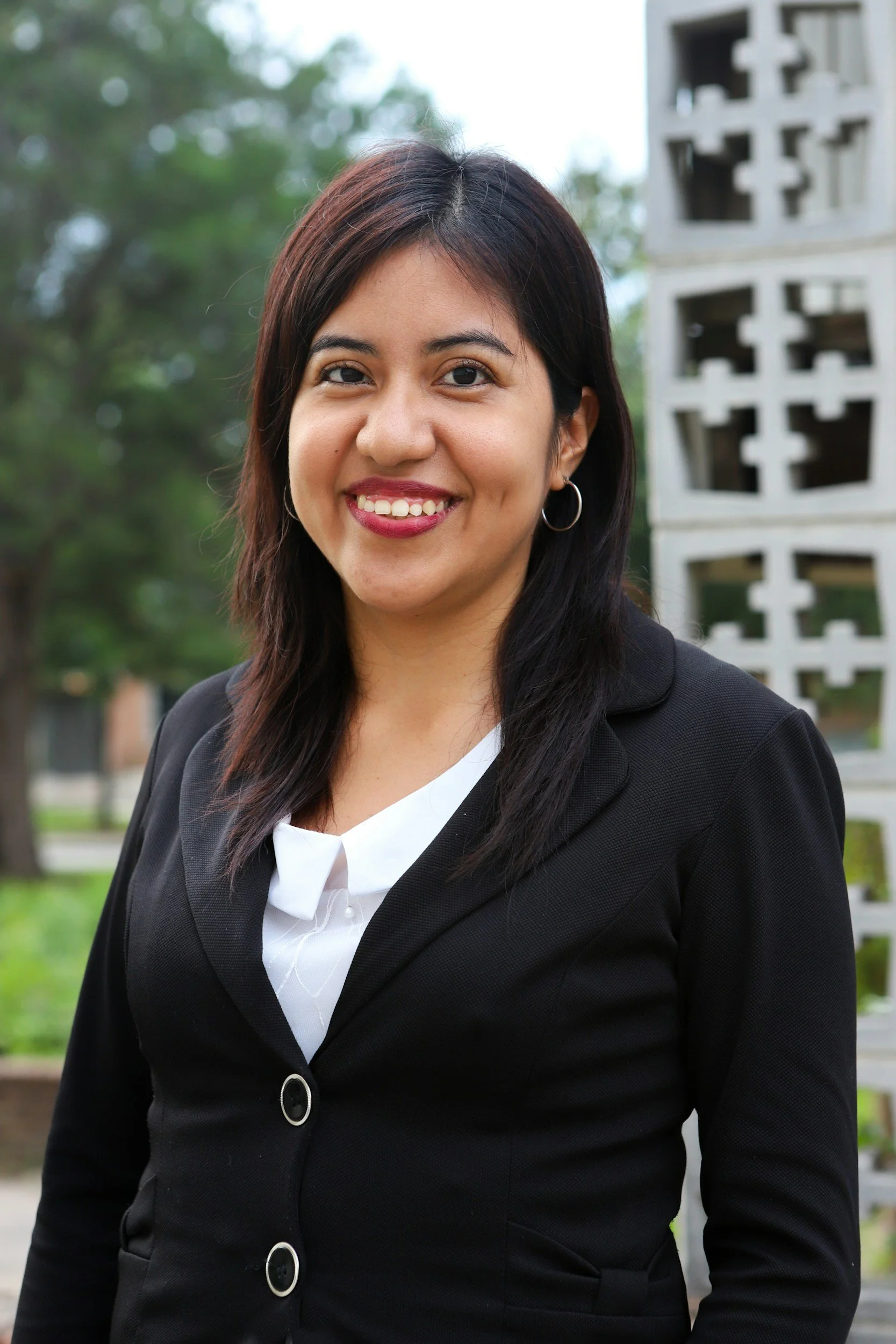 A woman outdoors wearing a black blazer and white blouse, smiling, with trees and abstract concrete sculpture in the background.