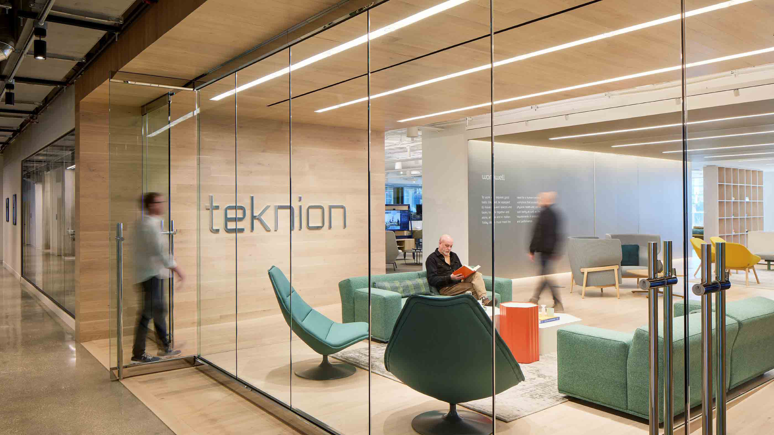 Modern office lobby with glass walls, green and yellow chairs, and a man sitting on a green couch reading a book.