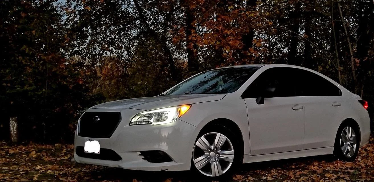 A white sedan car parked on a street at dusk with fallen leaves on the ground and trees in the background.