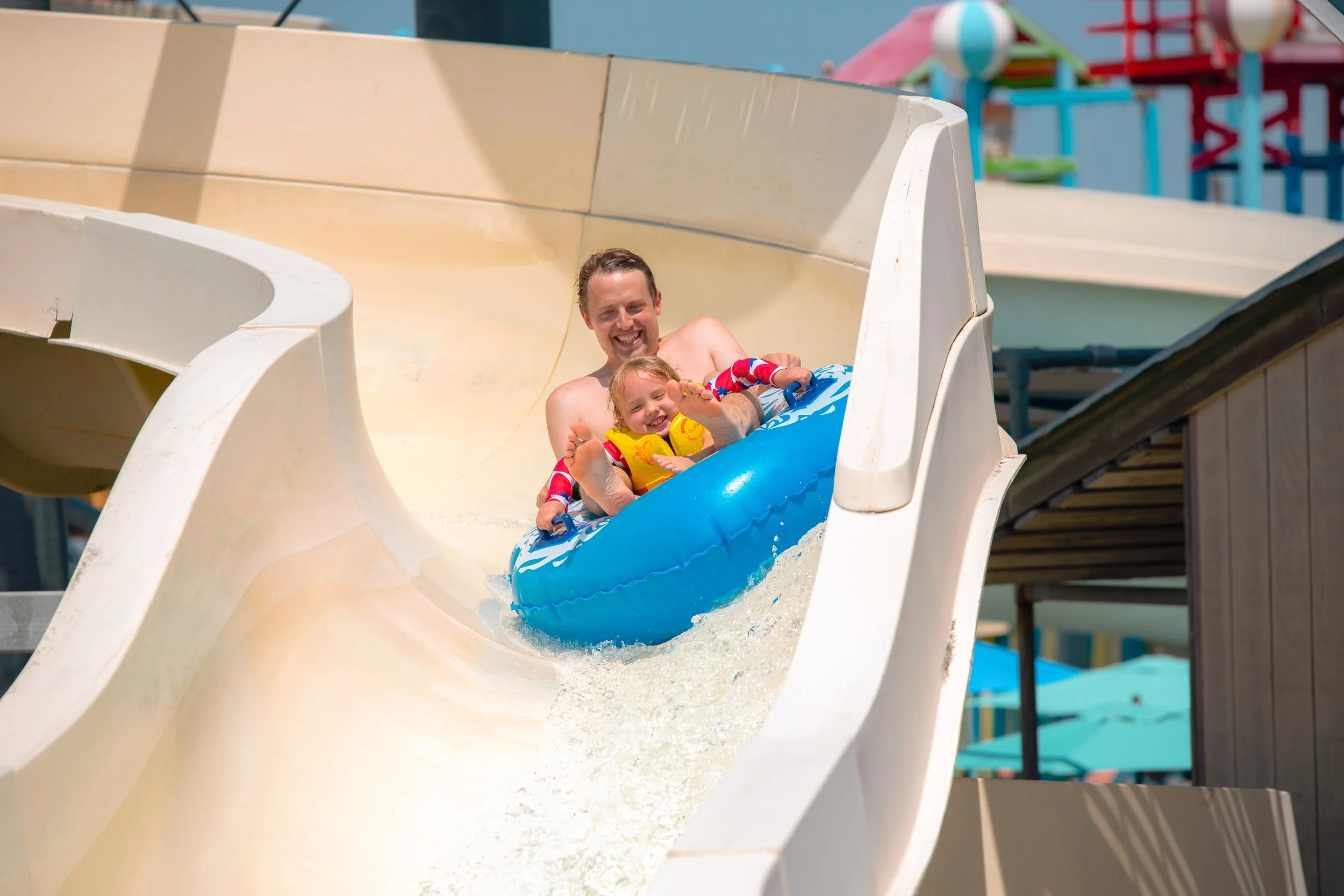 A man and a young girl enjoy riding down a water slide on an inflatable tube at a water park on a sunny day.