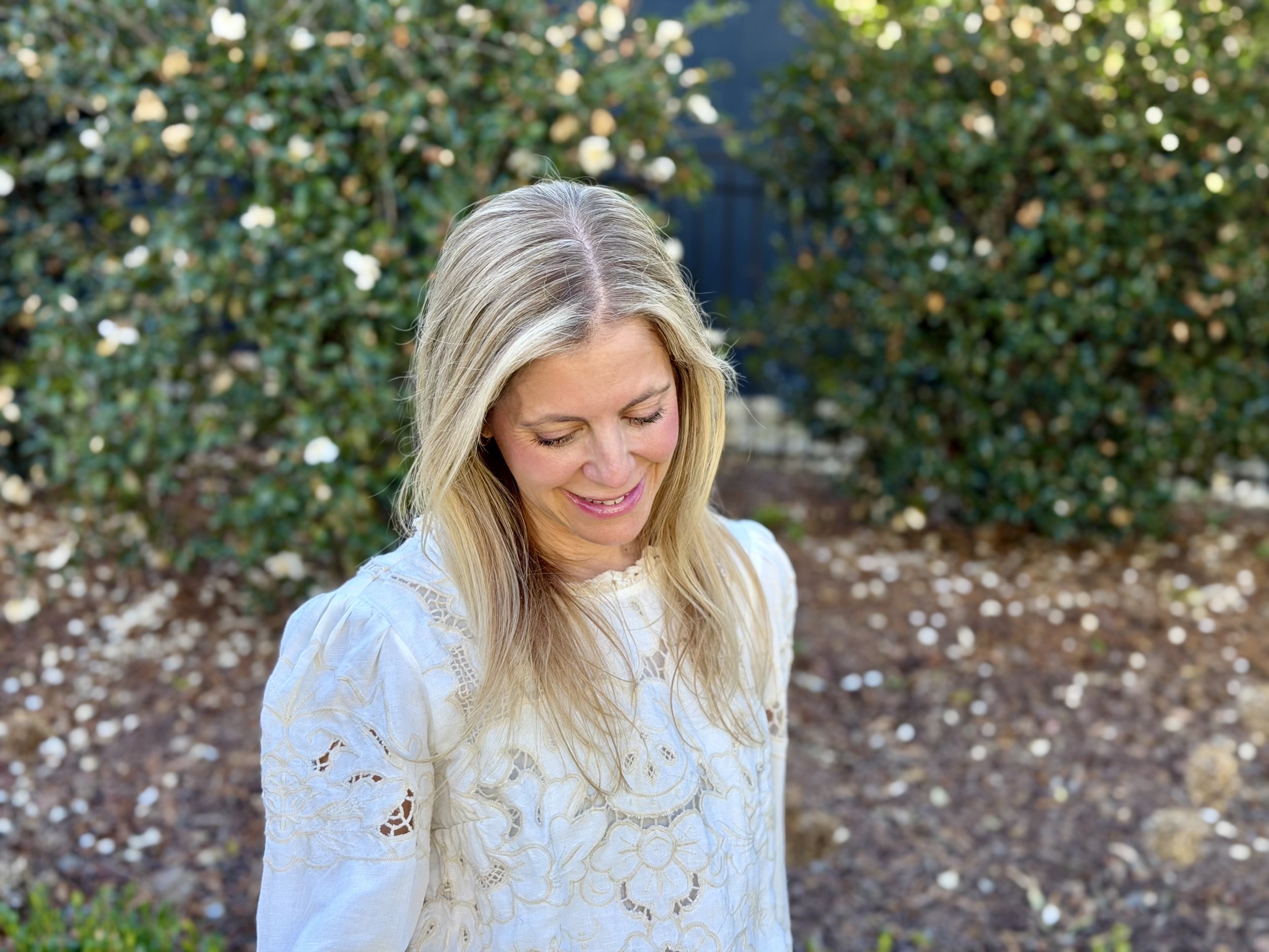 A woman with long blonde hair looking down and smiling outdoors, wearing a white lace blouse, with green bushes and a dark fence in the background.
