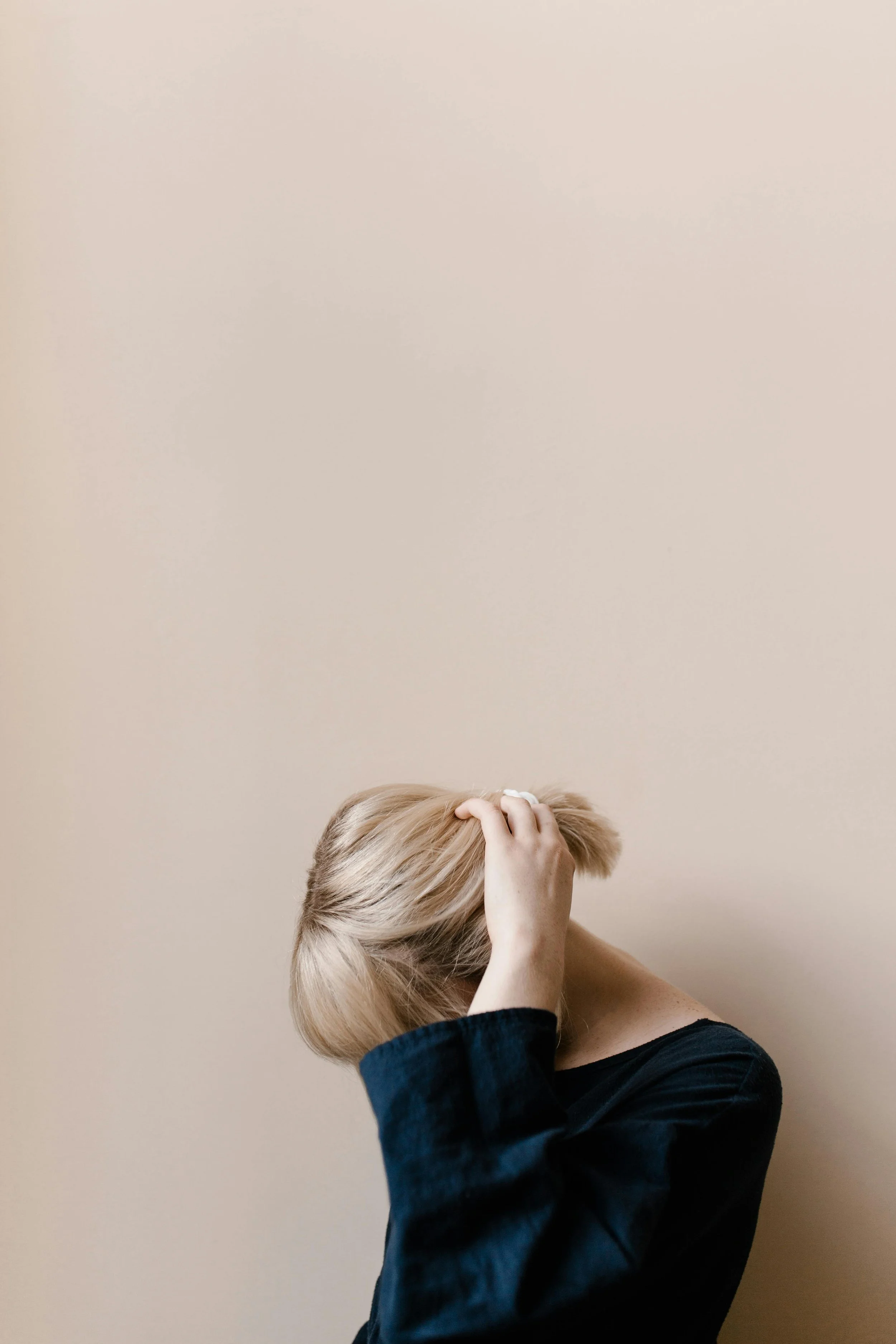 Person with blond hair, wearing a dark top, standing against a plain beige wall, holding their head with one hand.