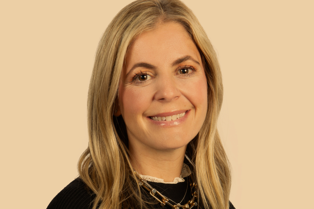 A woman with blonde hair smiling, wearing a black top and layered necklaces, against a beige background.