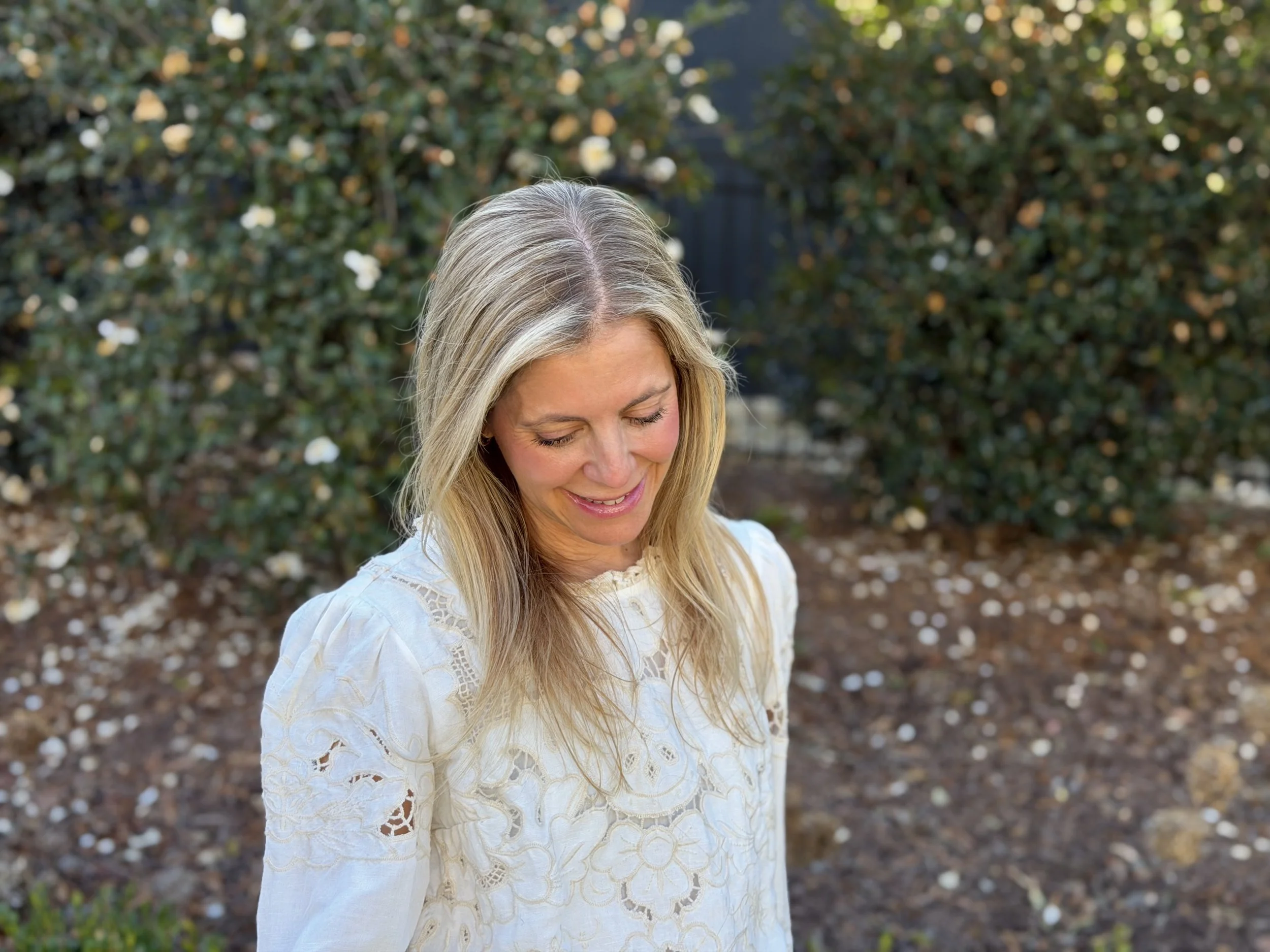 A woman with long blonde hair smiling and looking downward outside with bushes in the background.