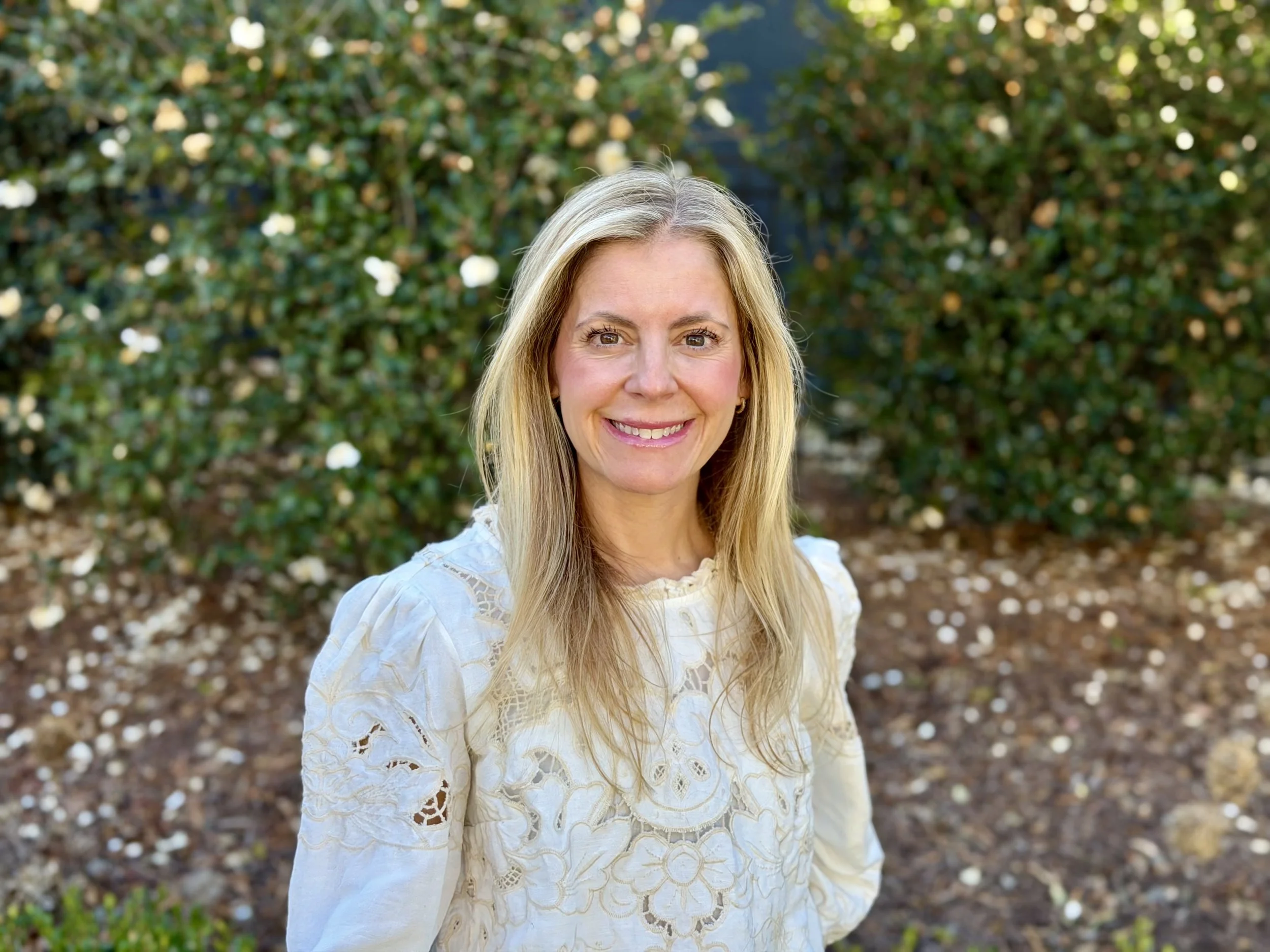 A smiling woman with long blonde hair, wearing a white lace blouse, standing outdoors in front of green bushes with small white flowers.