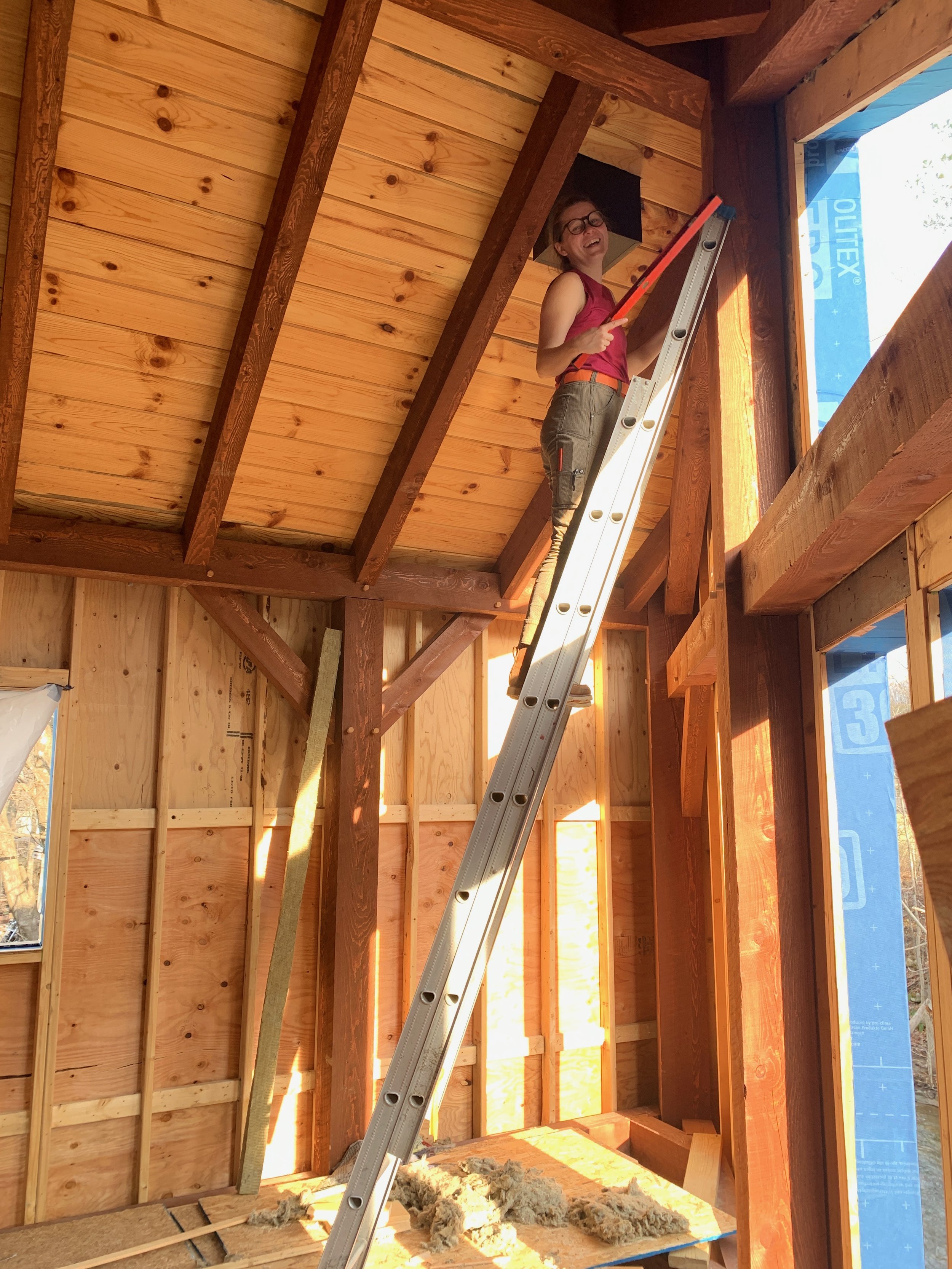 A woman standing on a ladder inside a room under construction, holding a level and smiling.