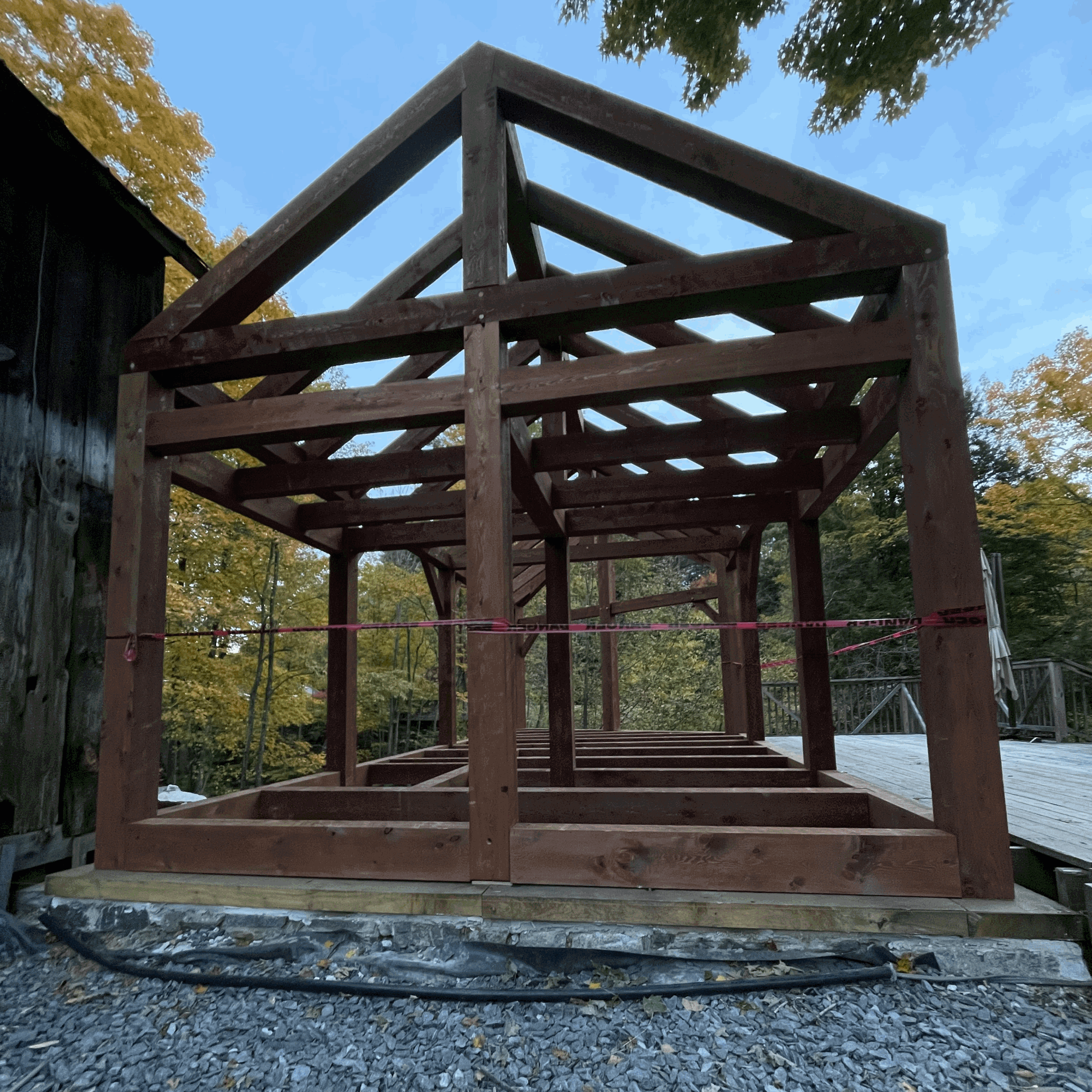Under construction wooden deck framework on a gravel foundation in an outdoor setting with trees and a blue sky.