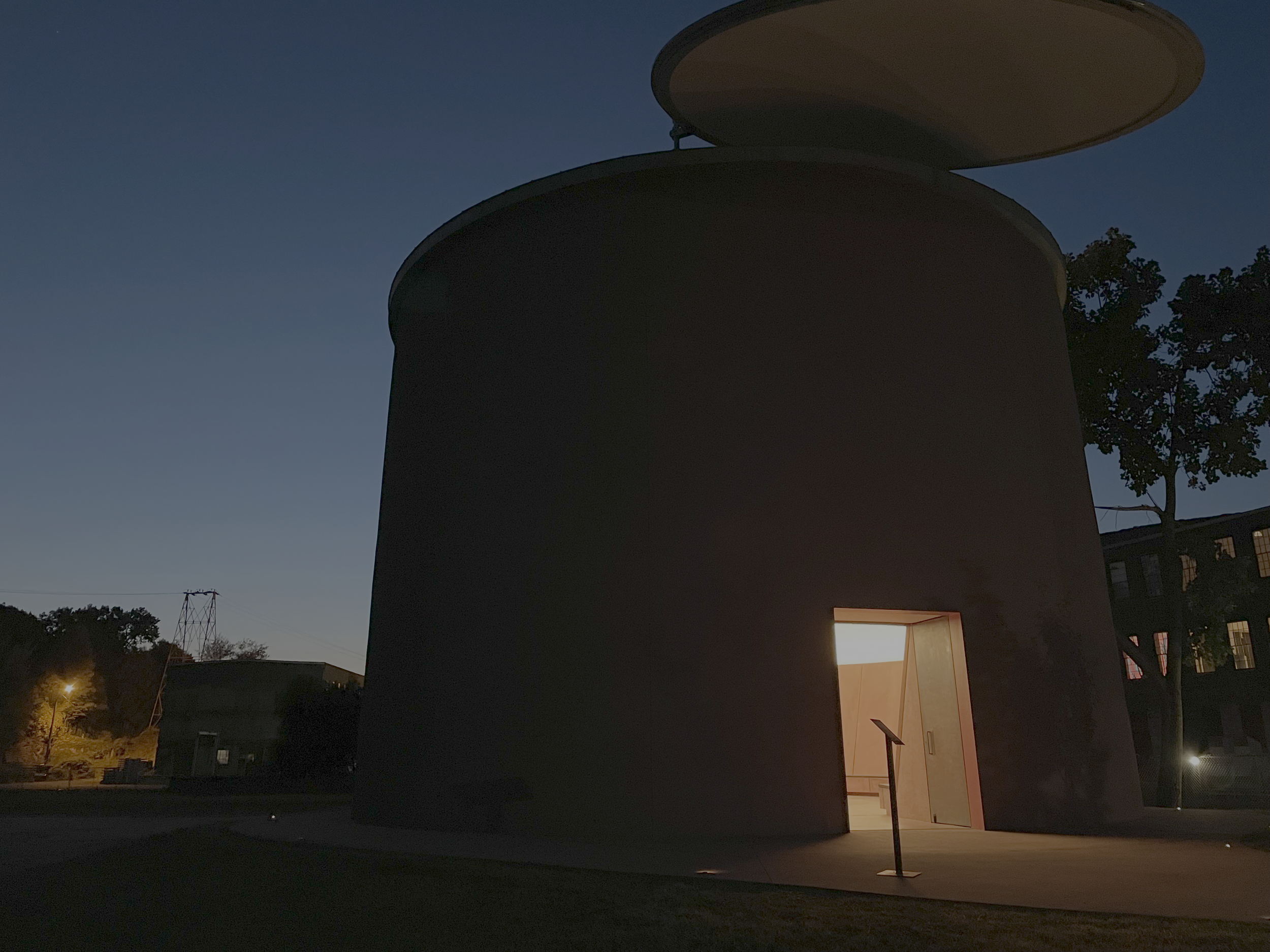 A modern, cylindrical building with a rounded roof, illuminated from within, during dusk or evening.