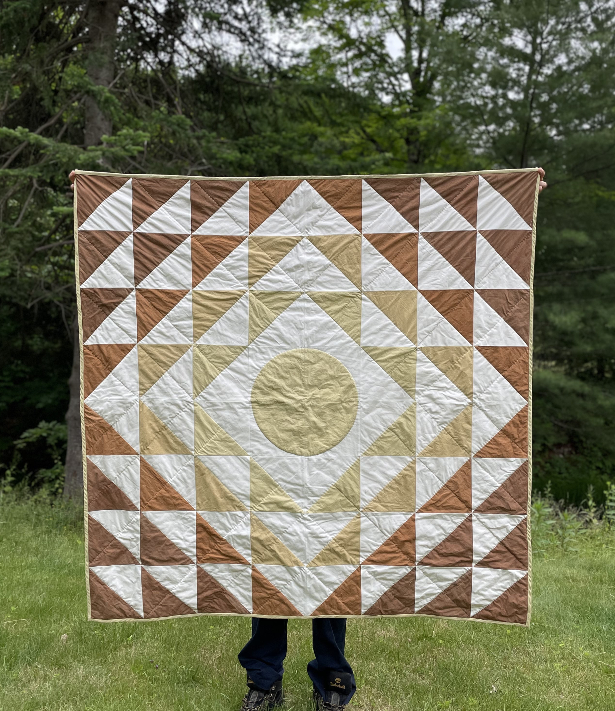 Person holding a quilt with a geometric pattern of white, beige, and brown triangles and circles outdoors on grass with trees in the background.