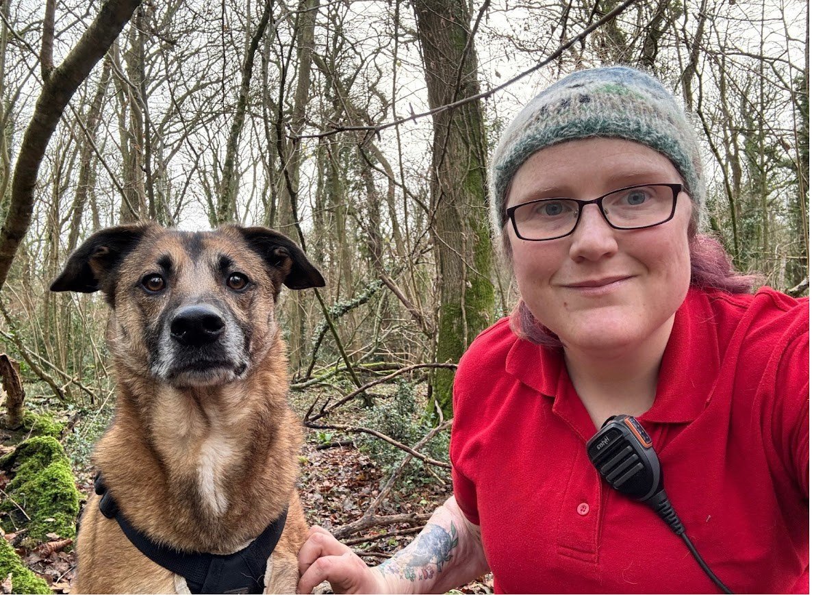 A woman with glasses, a gray knit beanie, and a red polo shirt, smiling, taking a selfie with a brown and black dog in a wooded area during winter or early spring.