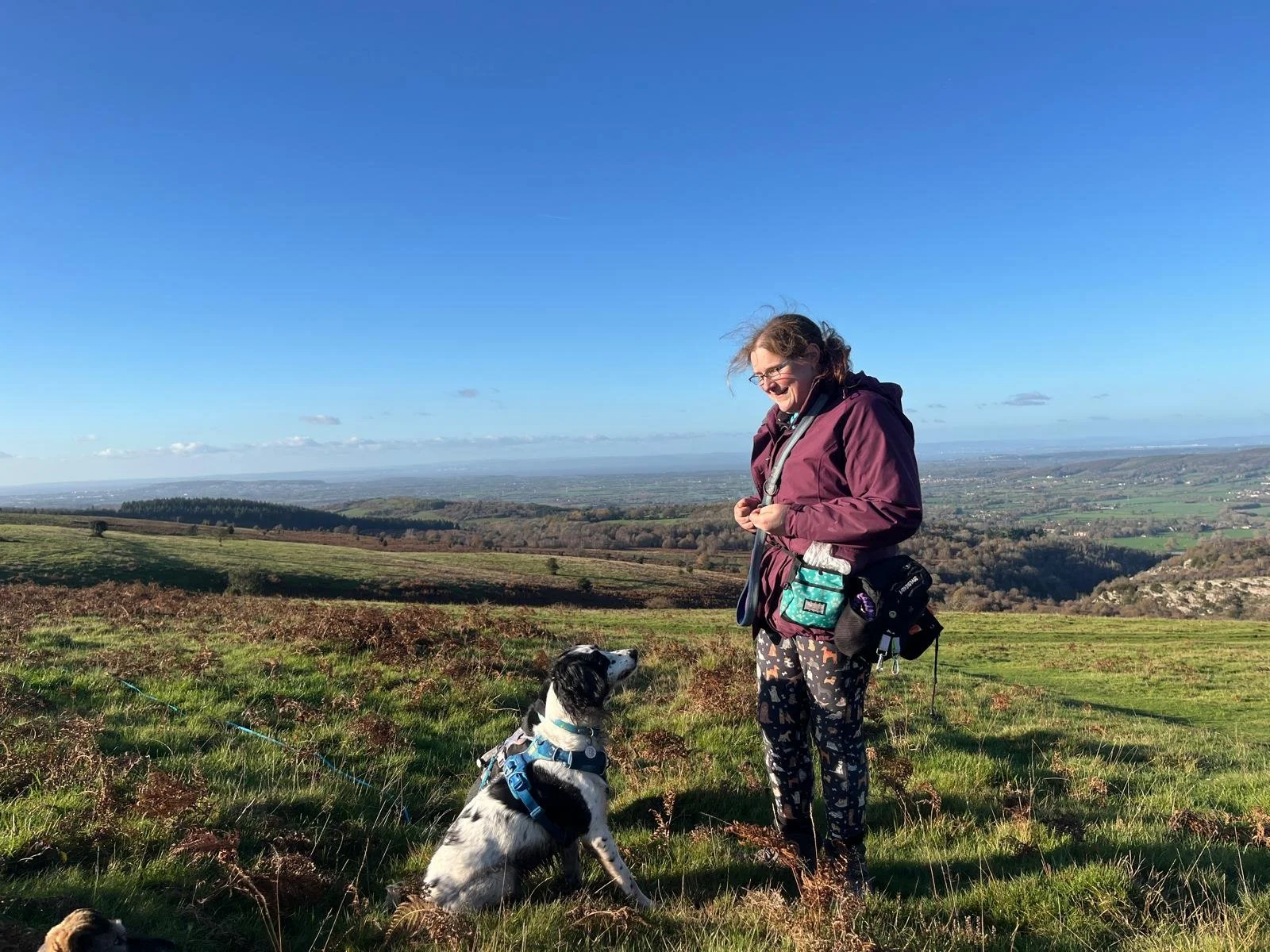 A woman with glasses and brown hair in a ponytail, wearing a maroon jacket and patterned pants, is standing outdoors on a grassy hill with a black and white dog sitting beside her. The woman is smiling and holding a leash, with a scenic landscape of rolling hills, fields, and a clear blue sky in the background.