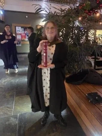 A woman dressed in a graduation gown holding a tall trophy or award, standing indoors near a decorated tree and large potted plants, with other people in the background.