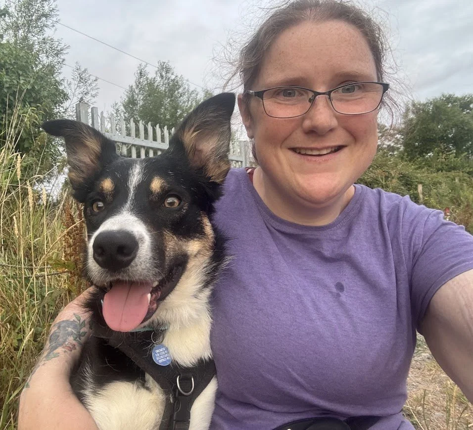 A woman with glasses and curly hair taking a selfie outdoors with a black and white dog, both smiling, in a natural setting with trees and bushes.