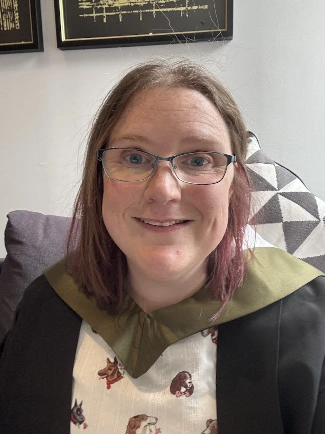 A woman with shoulder-length reddish-brown hair, wearing glasses, smiling at the camera. She wearing a graduation gown with a green hood