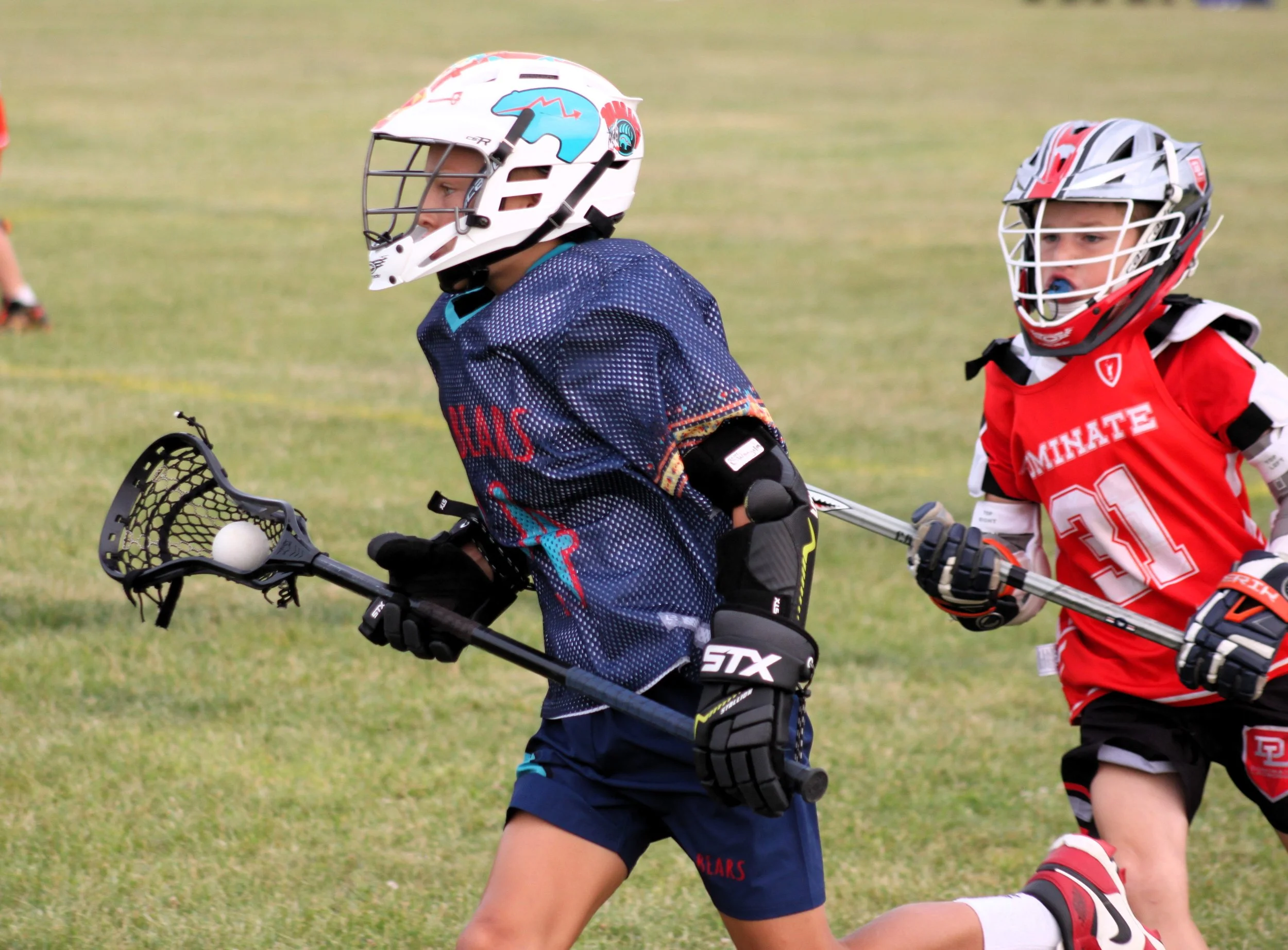 Two young boys playing lacrosse on a grassy field, wearing helmets, protective gear, and holding lacrosse sticks.