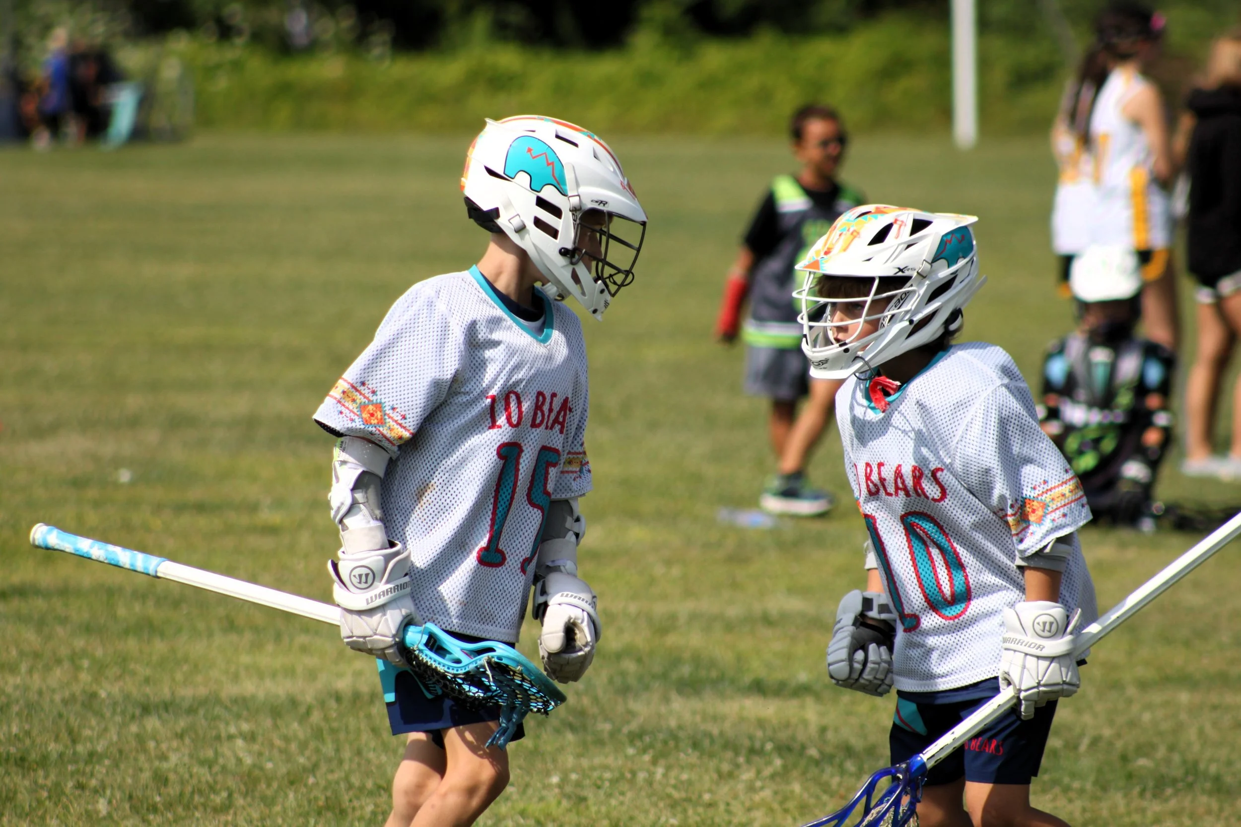Two young boys wearing protective lacrosse helmets and jerseys stand on a grassy field, holding lacrosse sticks and facing each other.