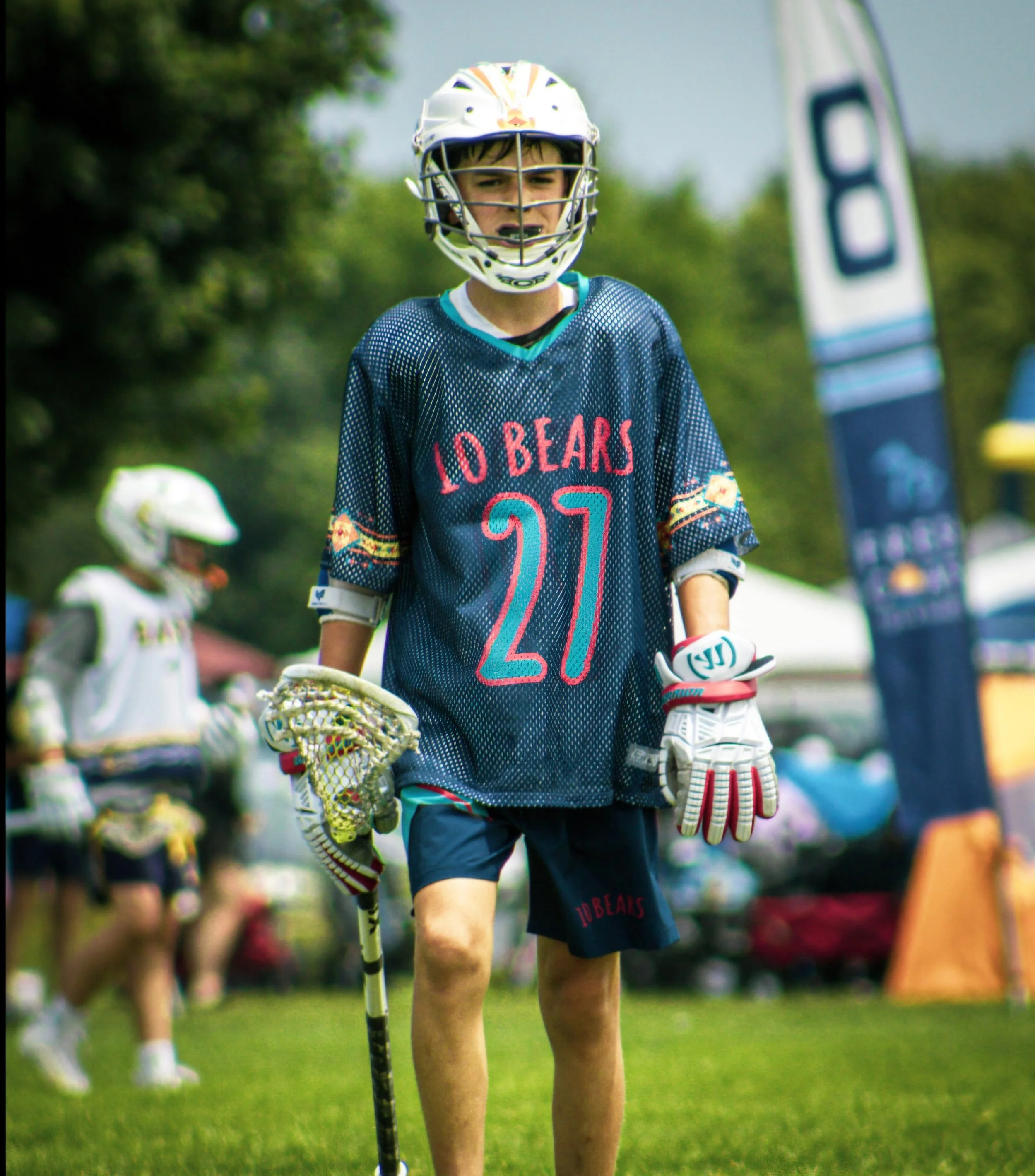 A young boy playing lacrosse outdoors on a grassy field, wearing a helmet and jersey, holding a lacrosse stick, and surrounded by other players and trees.