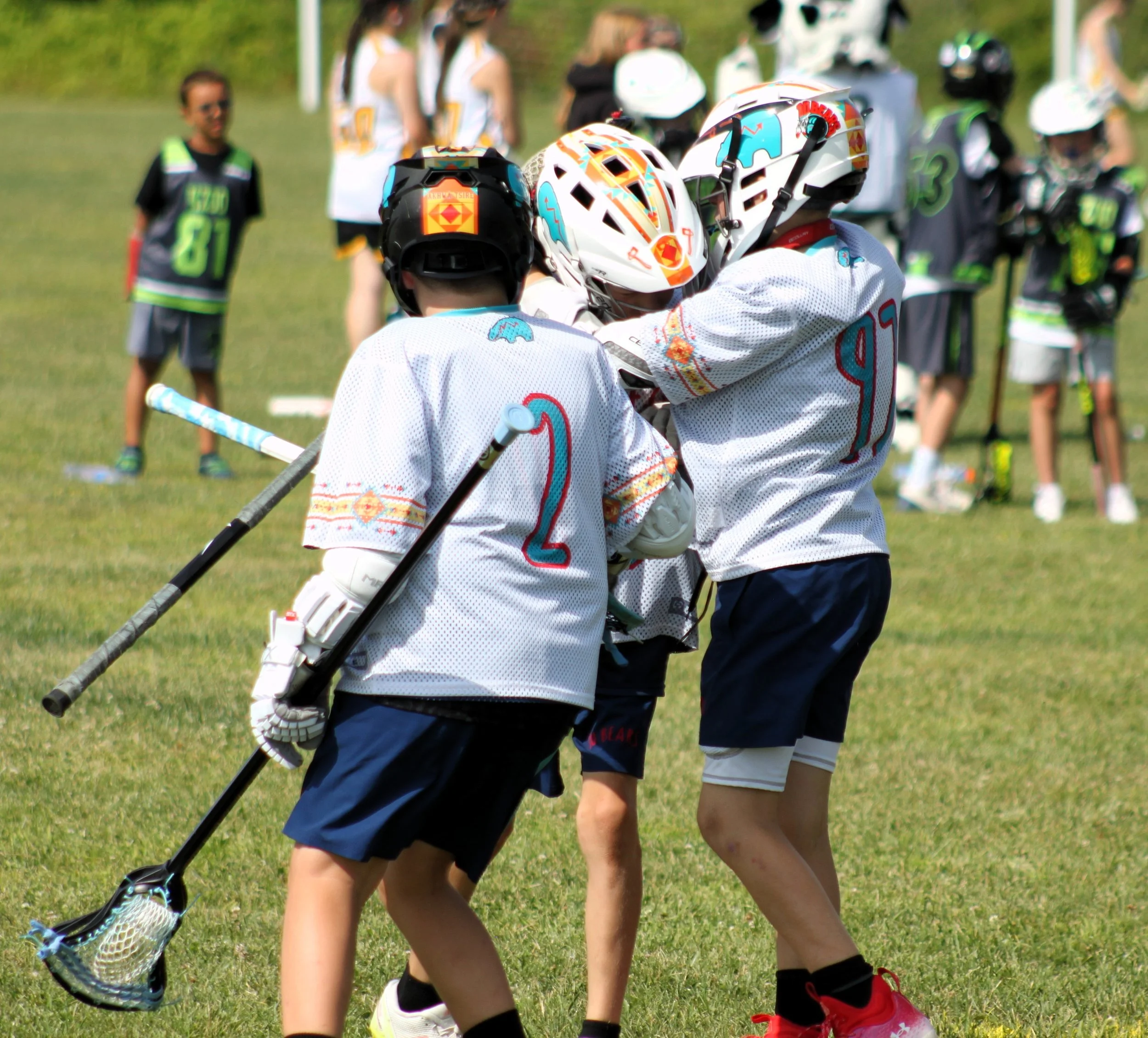 Two children wearing white hockey jerseys and helmets playing lacrosse outdoors on grass field, with adults and children in sports jerseys in the background.