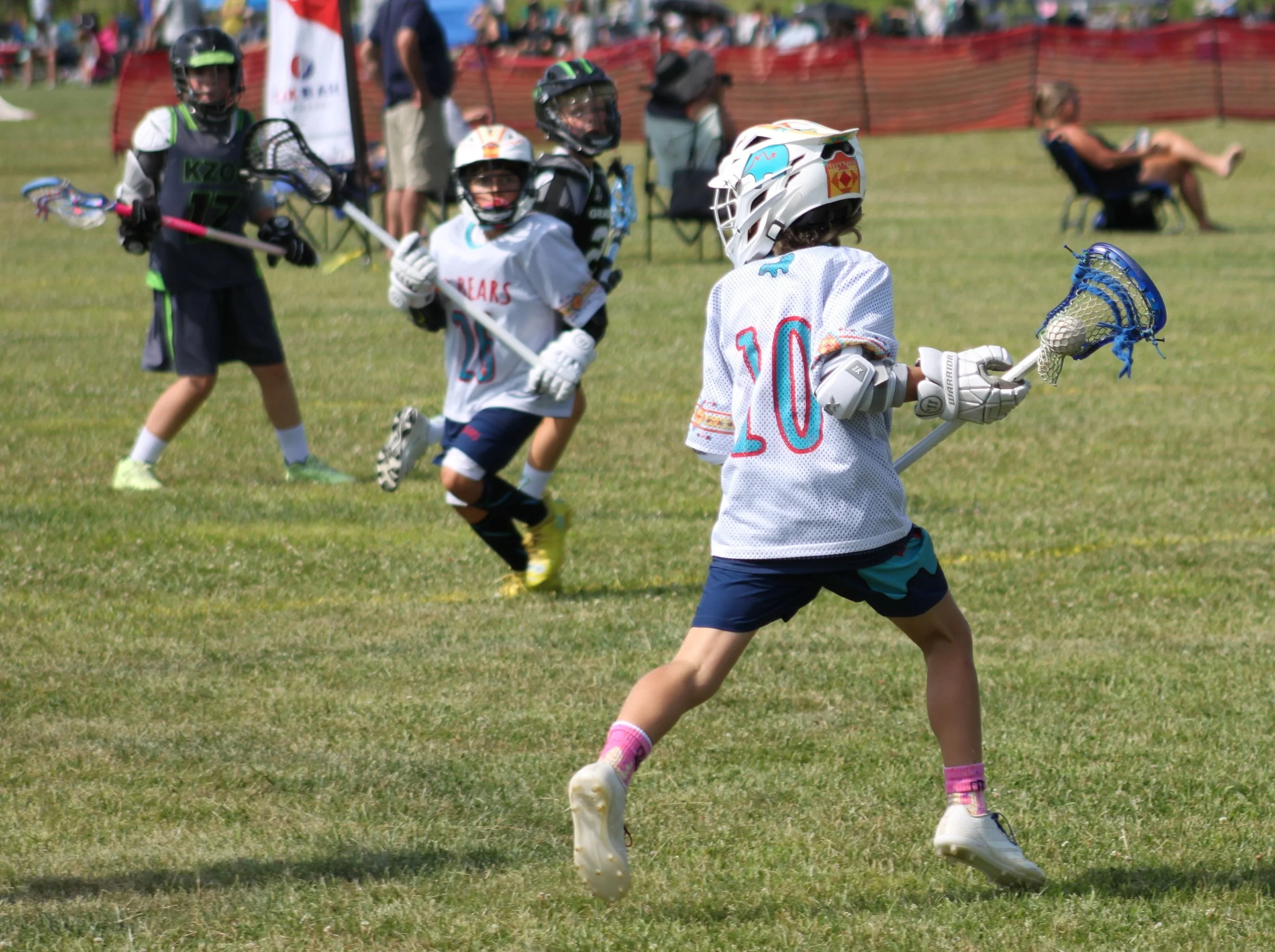 Children playing lacrosse on a grassy field during a sports event, with spectators in the background.