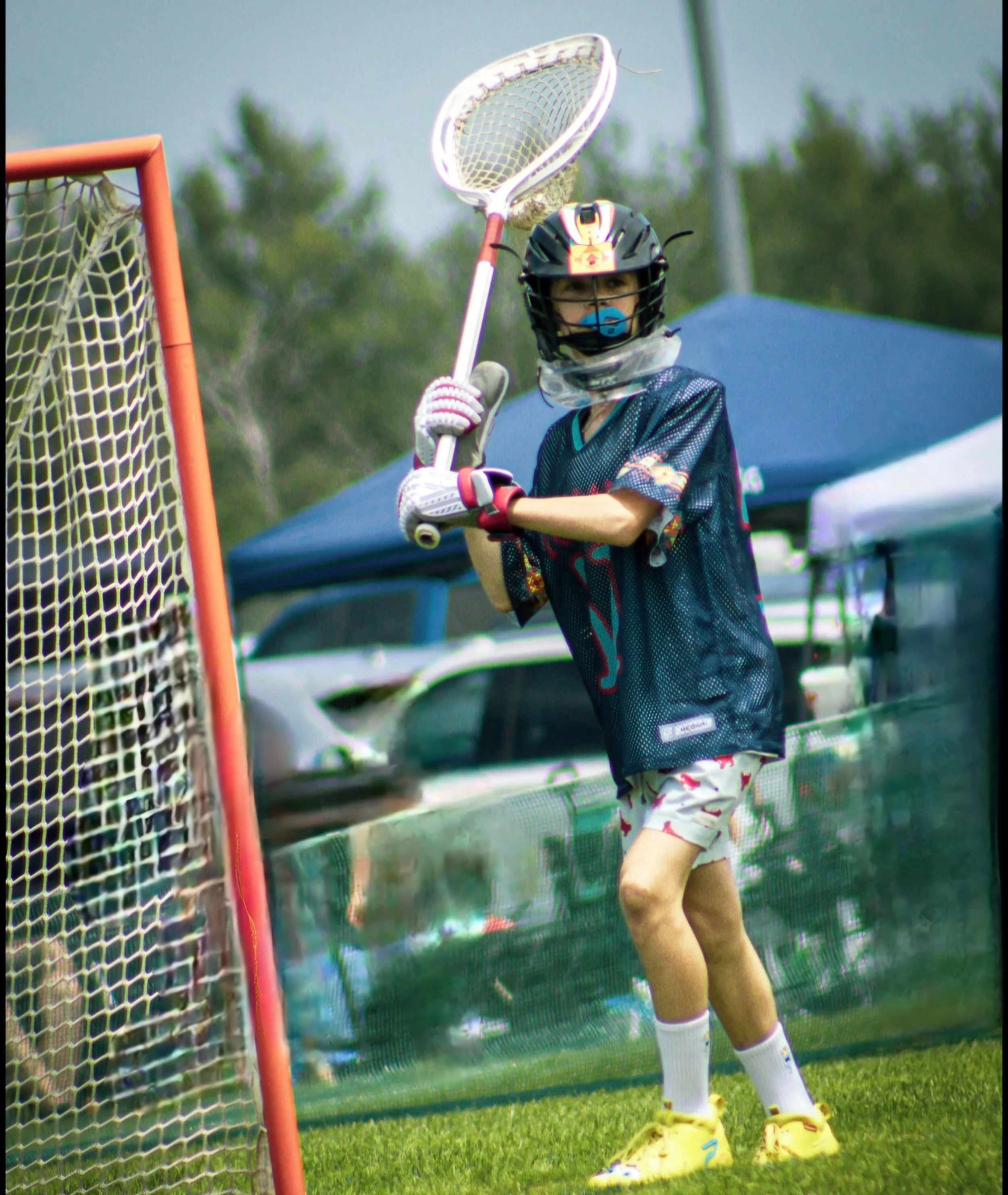 A young boy wearing a helmet, jersey, gloves, and yellow cleats, holding a lacrosse stick near a goal on a grassy field during daytime.