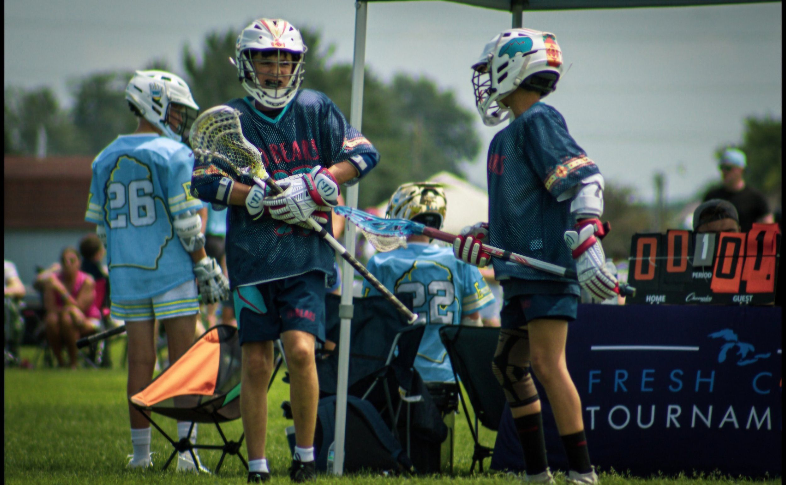 Children playing lacrosse on a grassy field, wearing helmets and sports gear, with a scoreboard and spectators in the background.