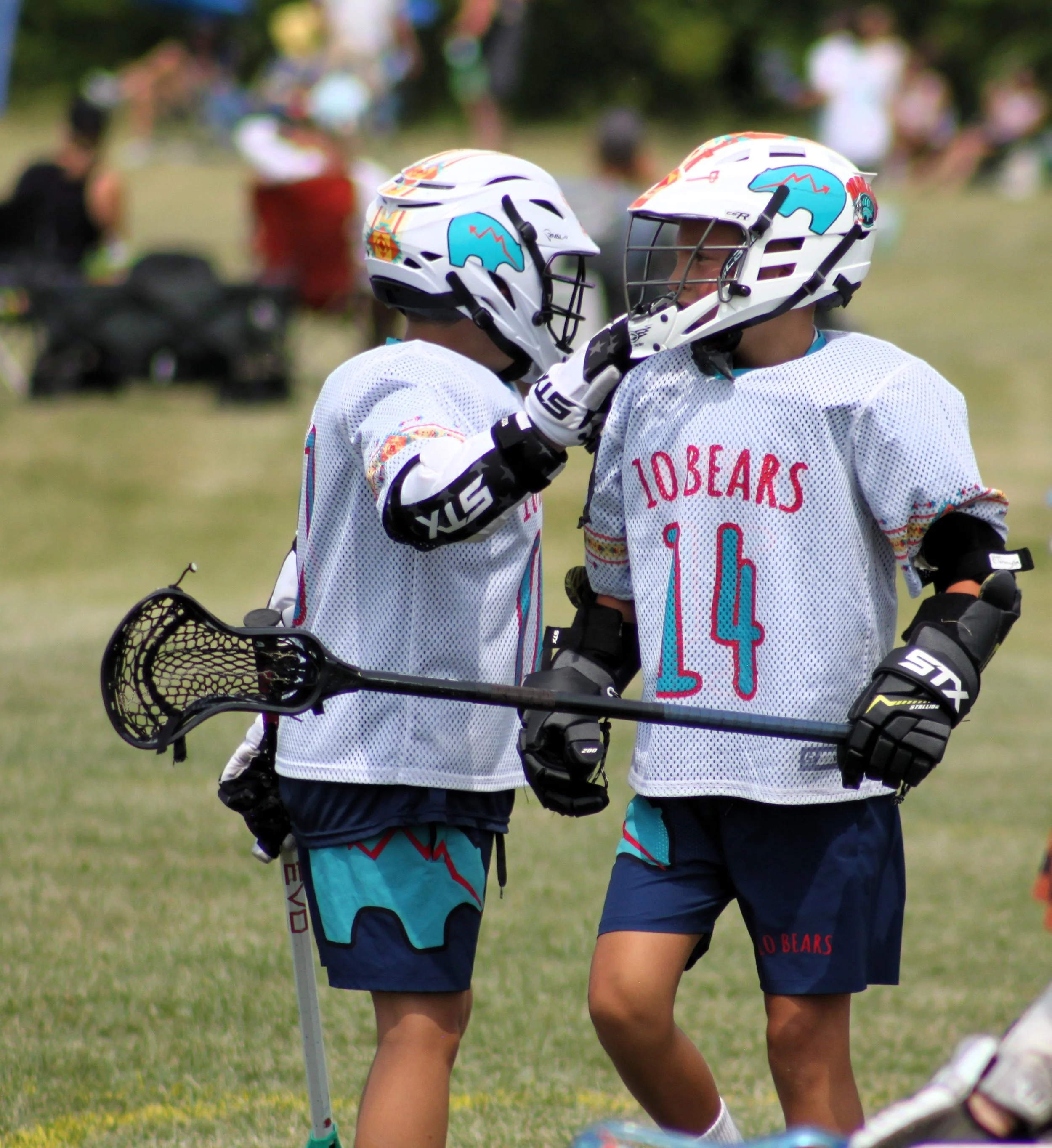 Two young female lacrosse players wearing white jerseys and helmets with blue and red graphic designs, standing close together and appearing to share a moment on the field, with a grassy background and blurred spectators.
