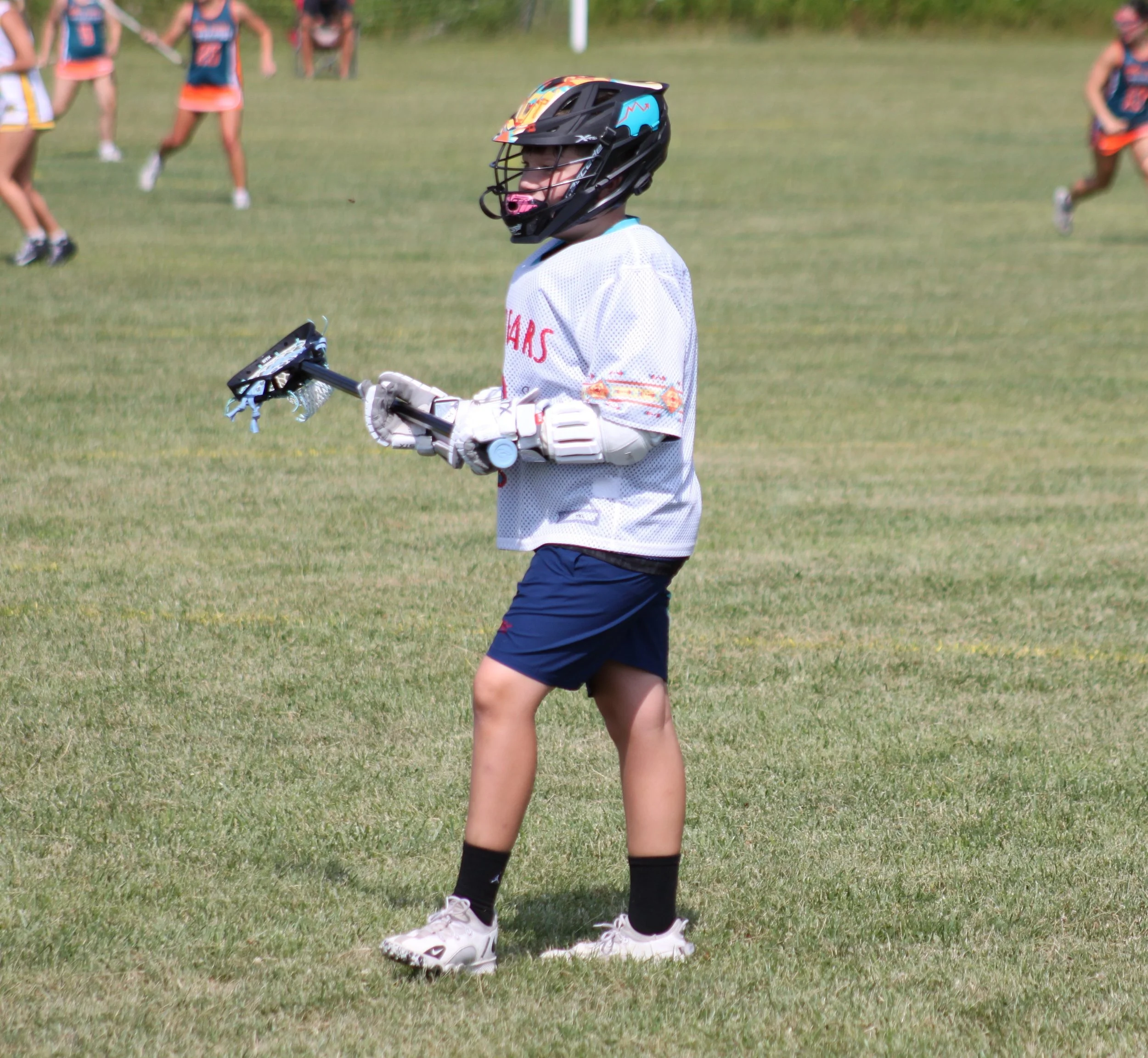 A young girl wearing a hockey helmet, white sports jersey, and black shorts walking on a grassy field during a sports game with other players in the background.