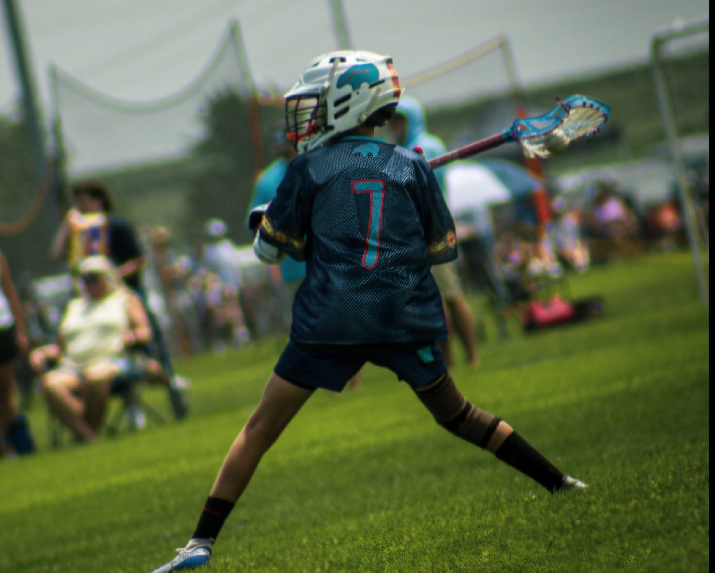 A youth wearing a white helmet with a blue dolphin design, black sports jersey with the number 7, and black shorts, is playing lacrosse on green grass, holding a lacrosse stick with a netted head.