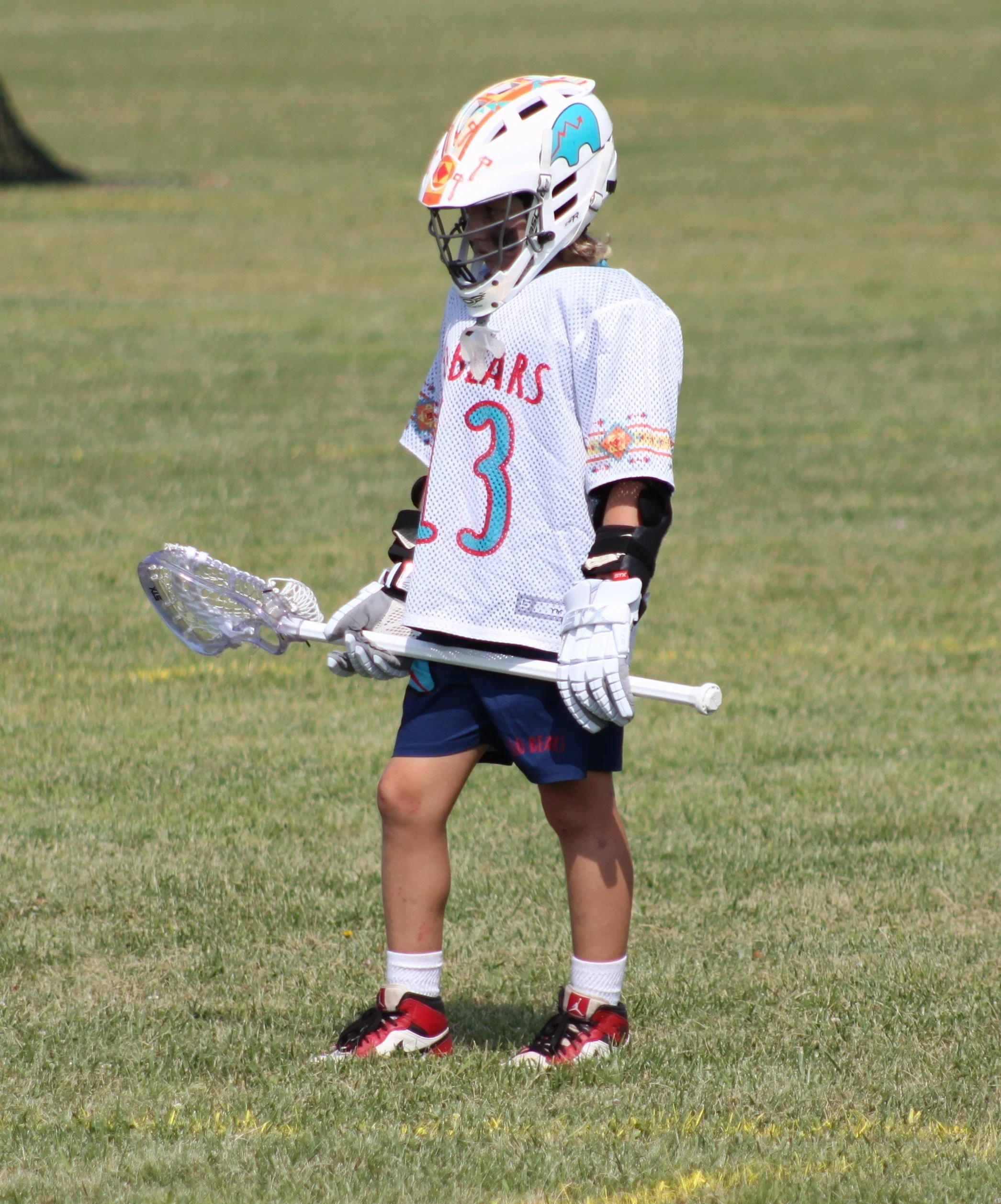 A young person wearing a helmet, hockey jersey, shorts, athletic shoes, and holding a lacrosse stick standing on a grassy field.