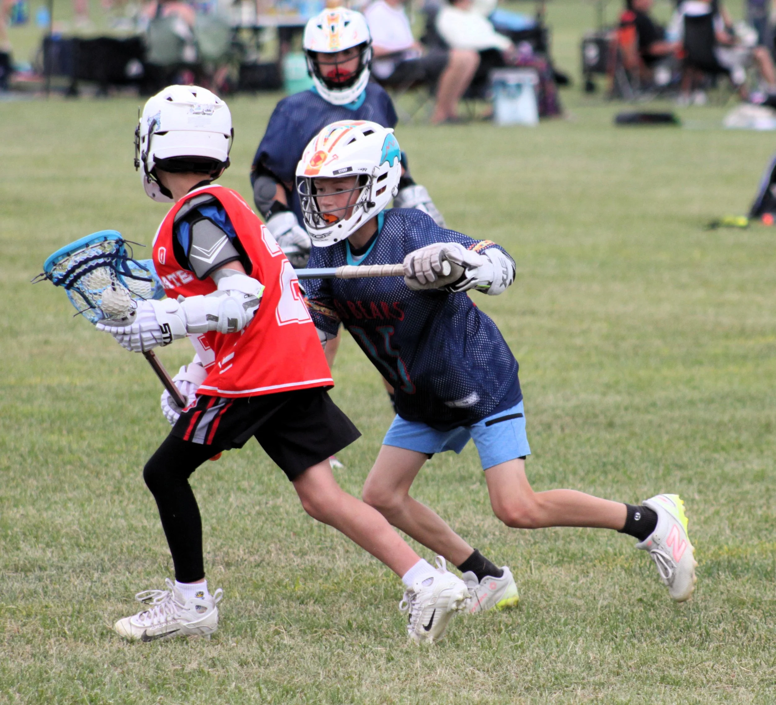 Two young boys wearing protective helmets and sports gear playing lacrosse on a grassy field, with other children and spectators in the background.