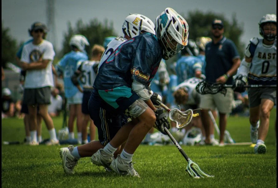 Lacrosse players on a field during a game, with one player in a dark jersey leaning forward to scoop the ball with a lacrosse stick