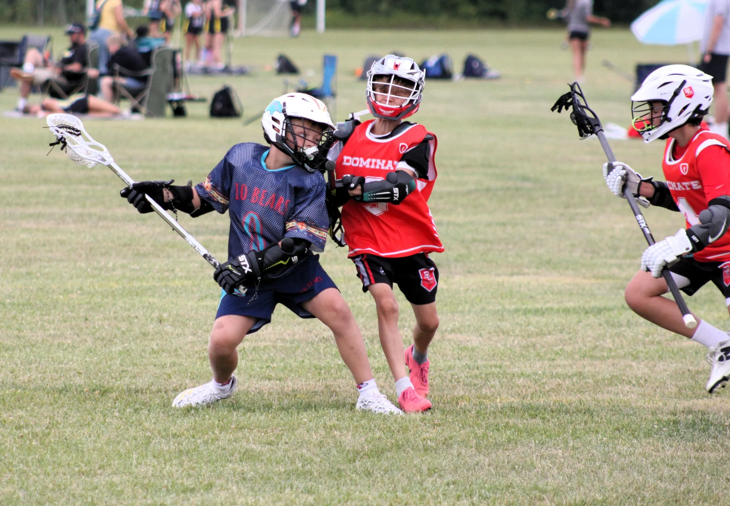 Three young lacrosse players wearing helmets, protective gear, and jerseys in red and blue are playing on a grassy field. One player in blue is holding a lacrosse stick, while two players in red are defending.