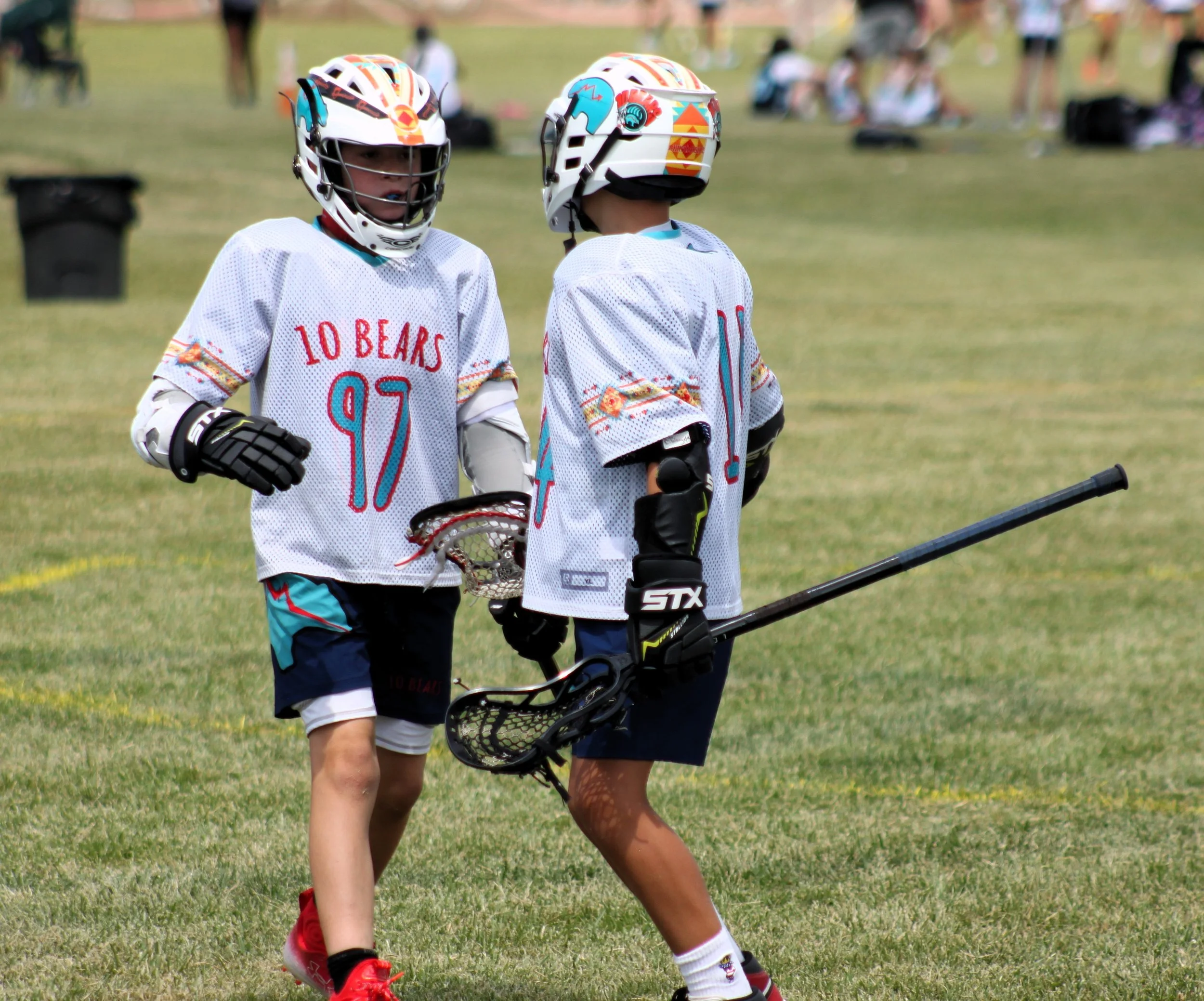 Two young kids wearing helmets and sports gear, walking on a grassy field, holding lacrosse sticks, during a youth sports game or practice.