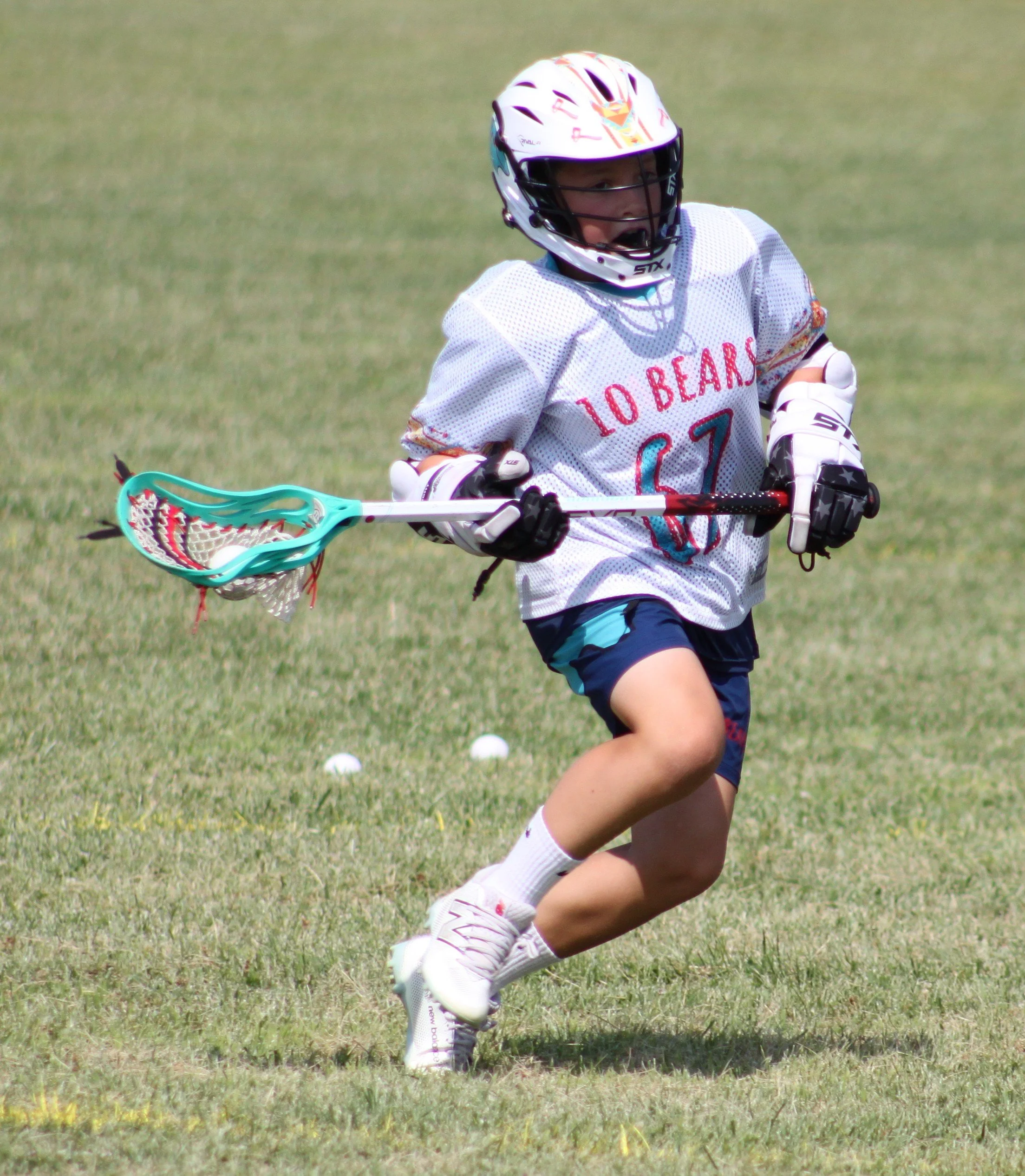 Young boy in a white sports jersey, wearing a helmet and gloves, running on a grassy field with a lacrosse stick in hand, chasing a small white ball.