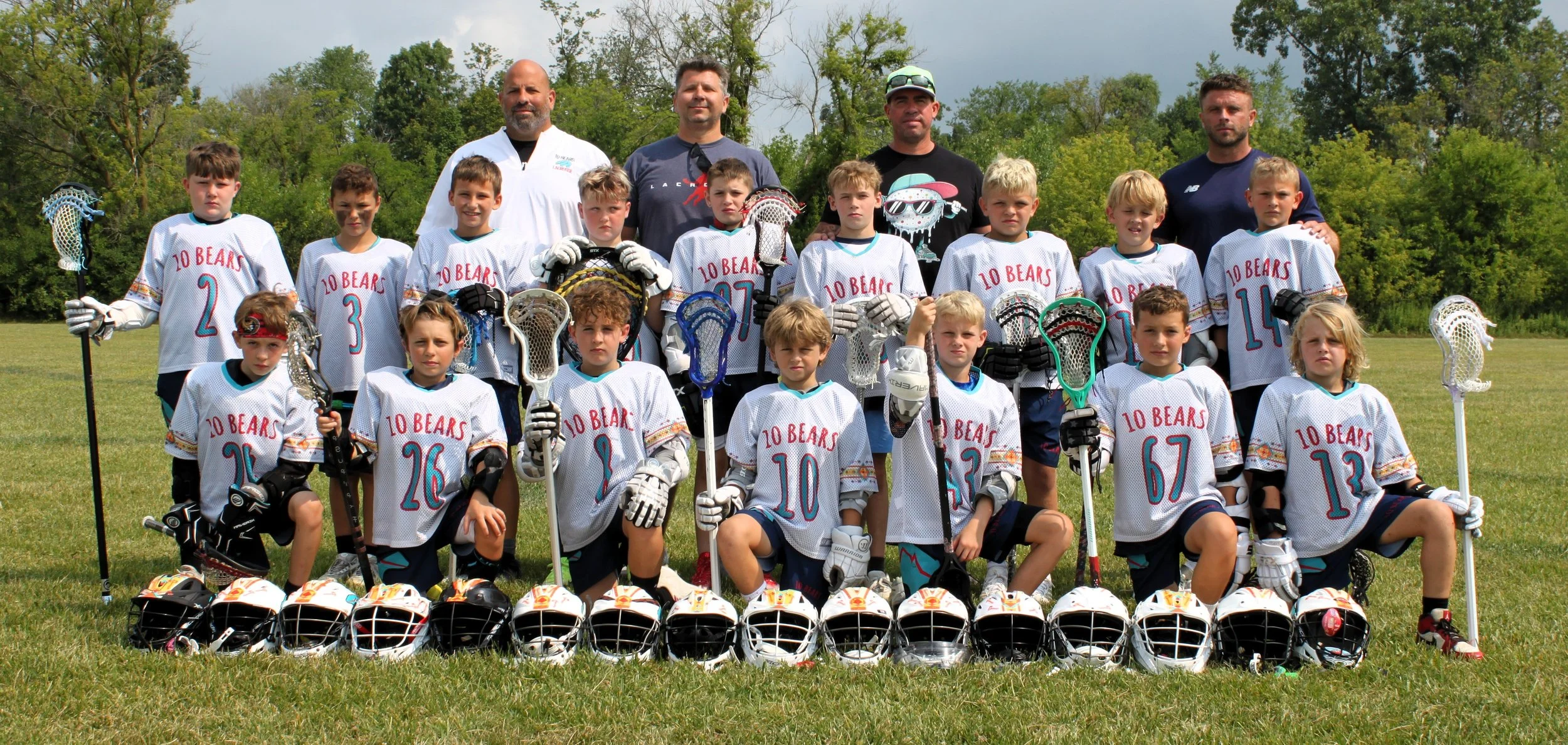 A youth lacrosse team called '10 Bears' posing for a group photo on a grassy field with coaches behind them. The team members are holding lacrosse sticks, and helmets are lined up in front of them. The team is wearing matching white jerseys with colorful details and various numbers.