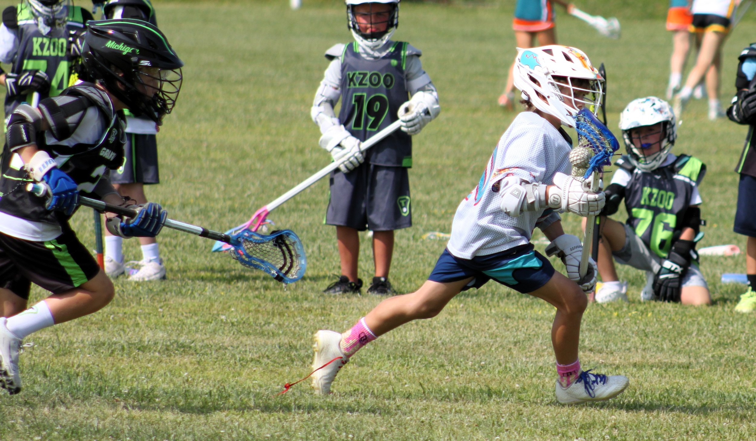 Young boys and girls playing lacrosse outdoors on a grassy field, wearing helmets and sports gear, with some kids sitting and watching in the background.