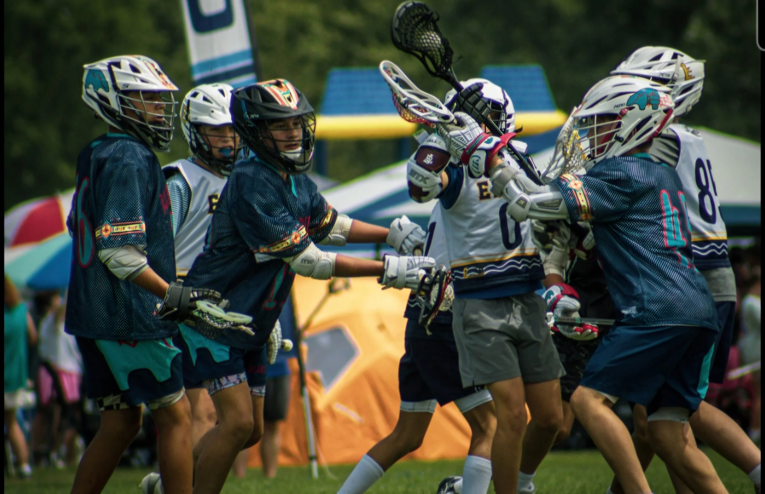 Lacrosse players on the field in a game, players wearing helmets and jerseys, some reaching for the ball, outdoors with tents and trees in the background.