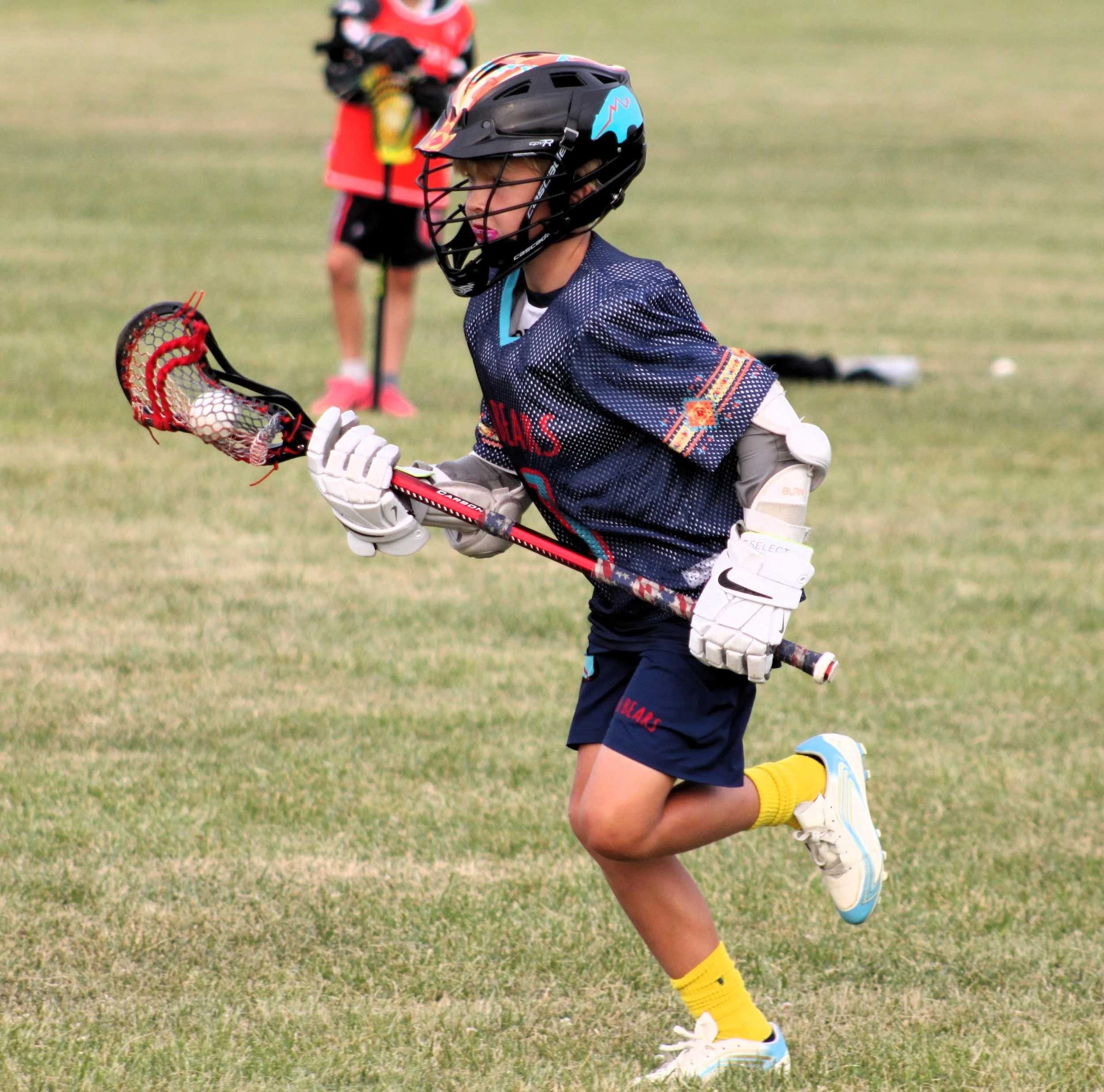 A young girl playing lacrosse on a grassy field, wearing a black helmet with colorful designs, protective gloves, a sports jersey, shorts, and athletic shoes, holding a lacrosse stick with a ball in the net.