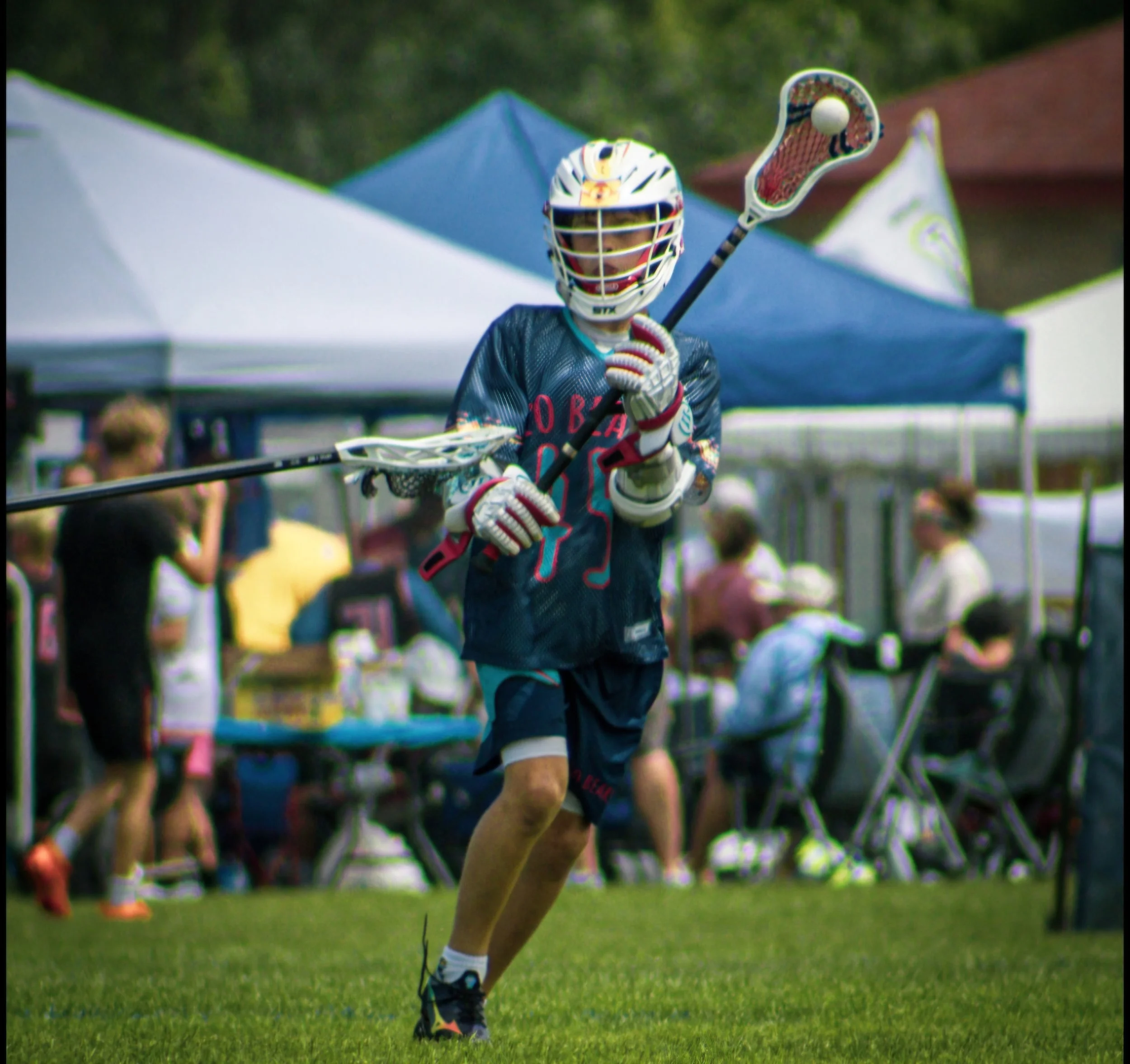 Lacrosse player wearing protective helmet and uniform, holding a lacrosse stick, running on a grassy field during a game with tents and spectators in the background.